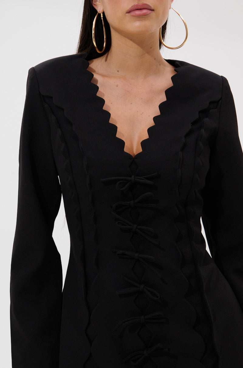 A woman models the CUTE AS CAN BE MINI DRESS WITH BOWS, a black tailored A-line mini dress with zigzag edges and decorative bows, paired with large hoop earrings. Her long dark hair frames her partially visible face and upper torso.