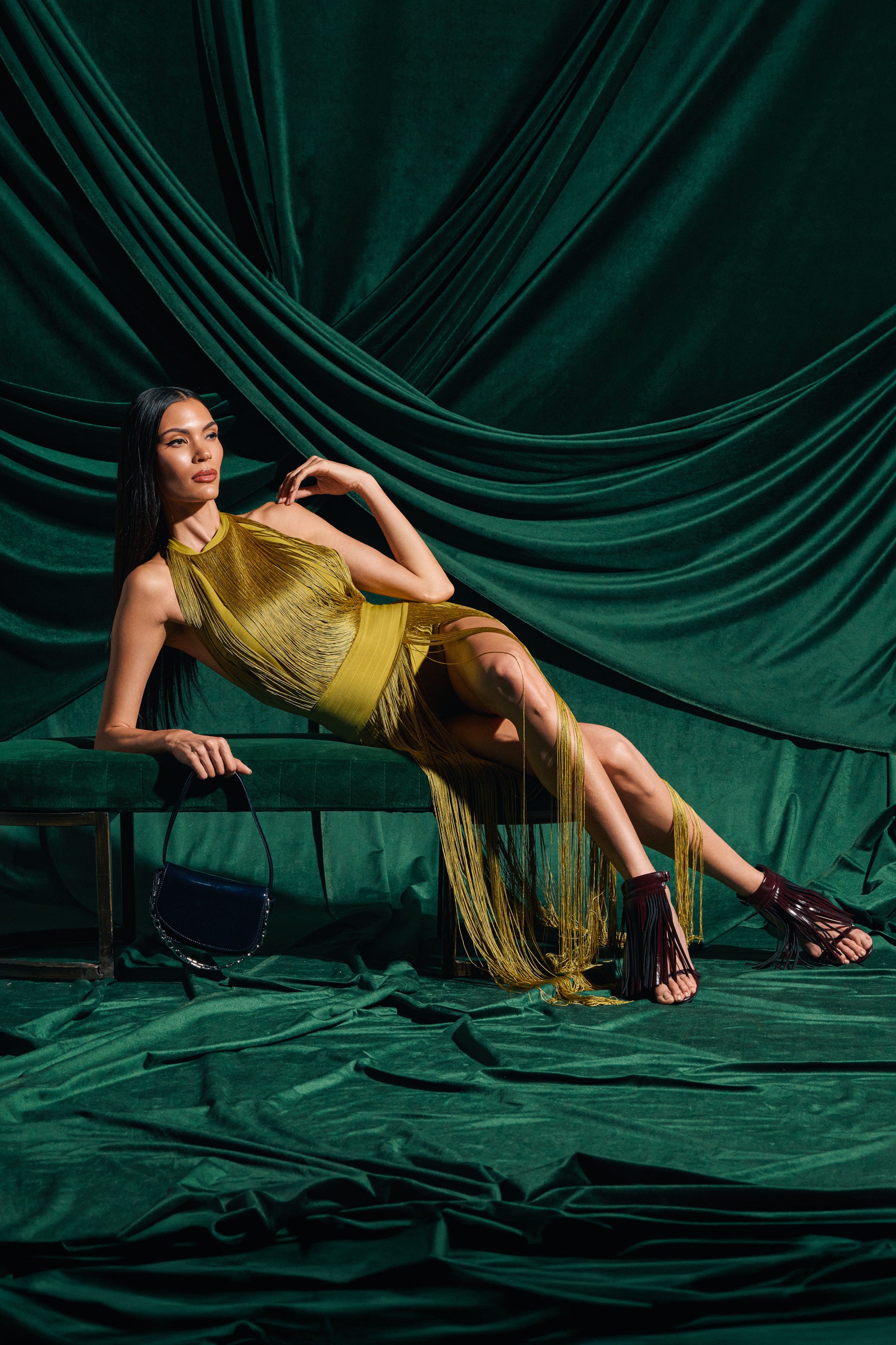 A woman in the RENEE FRINGE ROMPER IN GREEN lounges on a green velvet bench, surrounded by matching drapes. She holds a handbag, resting one arm on the bench, while fringe sandals complete her look.