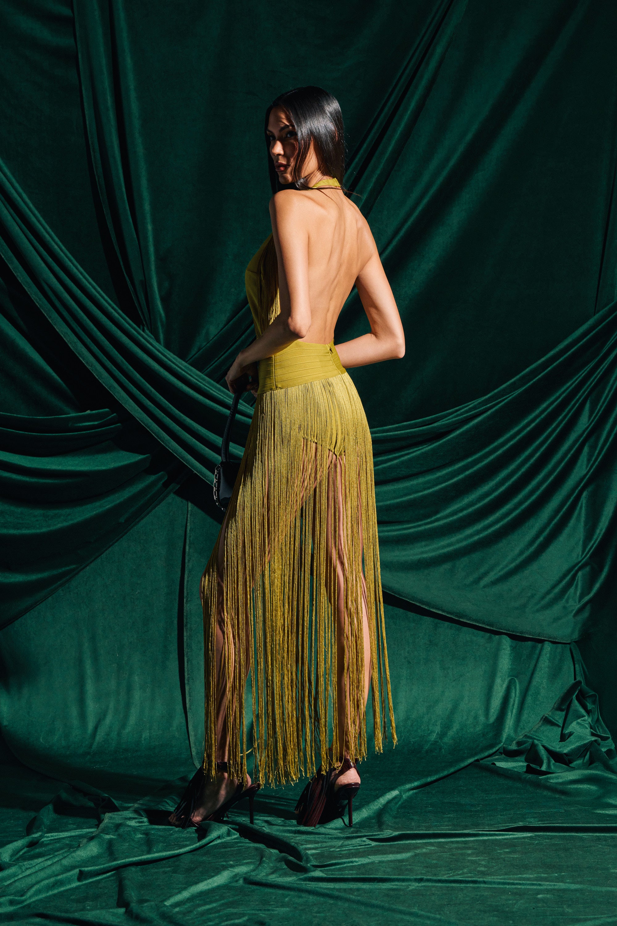 A woman with long dark hair poses in front of green draped fabric, wearing the RENEE FRINGE ROMPER IN GREEN—a backless, halter-neck bandage romper featuring a sheer, fringed skirt. She stands turned away, looking over her shoulder.