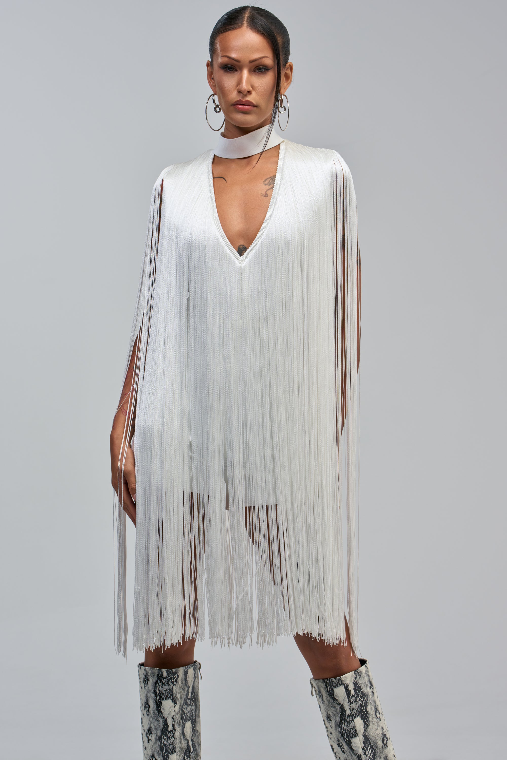 A woman stands confidently against a gray background in the SHAKE IT FOR ME SLEEVELESS BANDAGE ROMPER WITH FRINGE IN WHITE, styled with a matching choker and snakeskin knee-high boots.