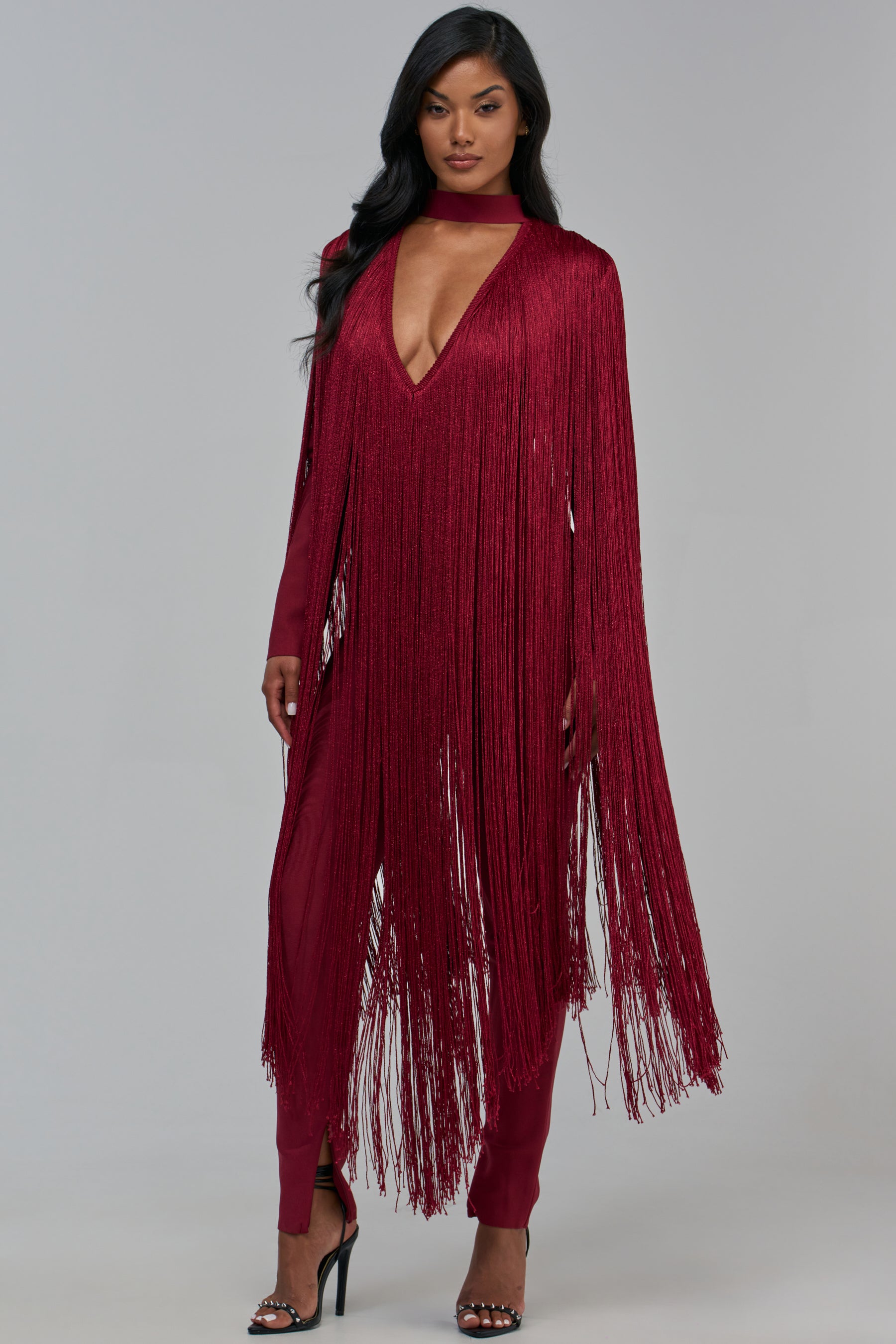 A woman wears the SHAKE IT FOR ME LONG SLEEVE BANDAGE JUMPSUIT IN BURGUNDY, featuring a deep V-neck and a floor-length fringe overlay.