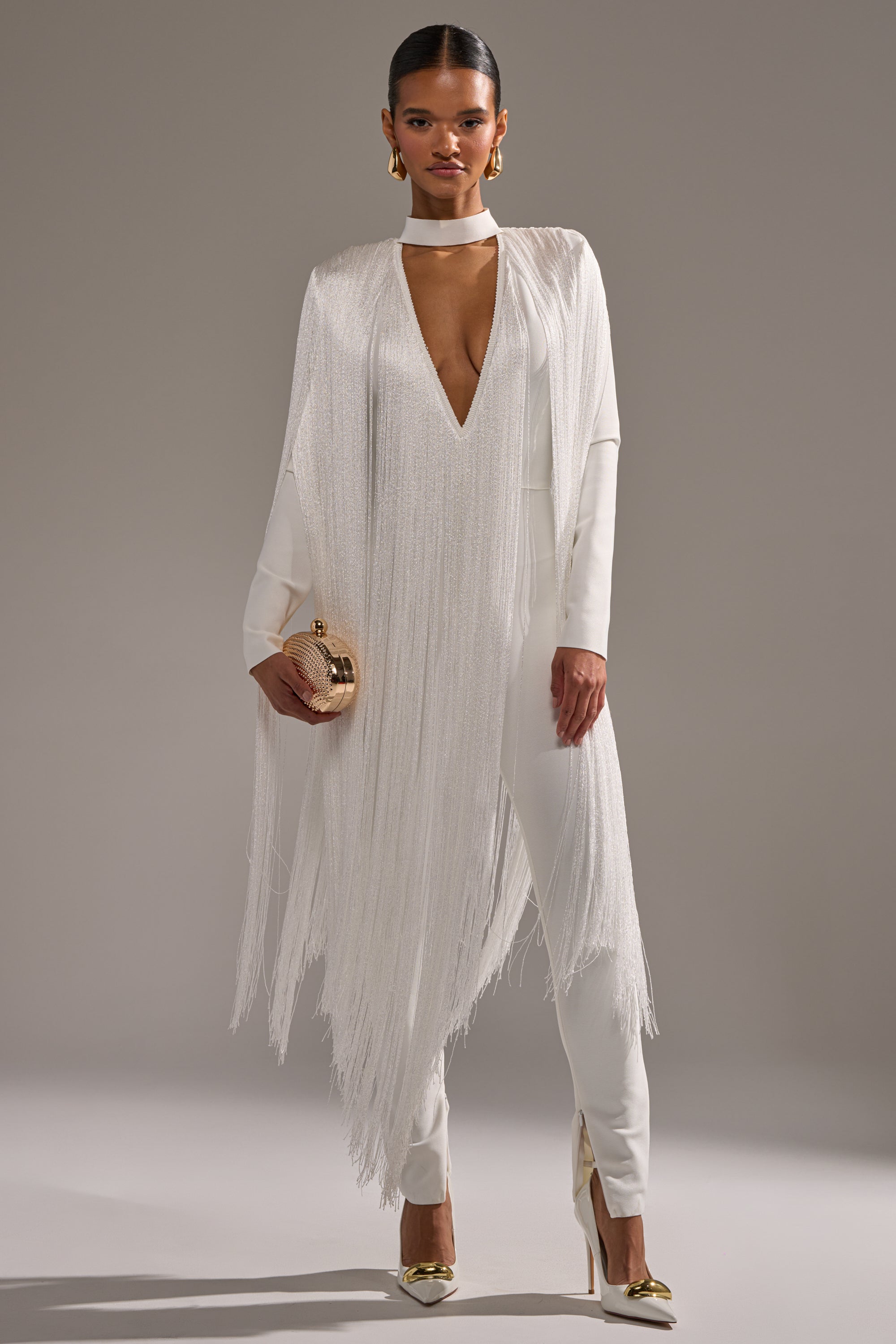 A woman stands confidently in the BELLE BANDAGE JUMPSUIT WITH FRINGE IN WHITE, featuring a deep V-neck and matching choker. She pairs it with gold heels, earrings, a gold clutch, and sleeked-back hair for a chic look.