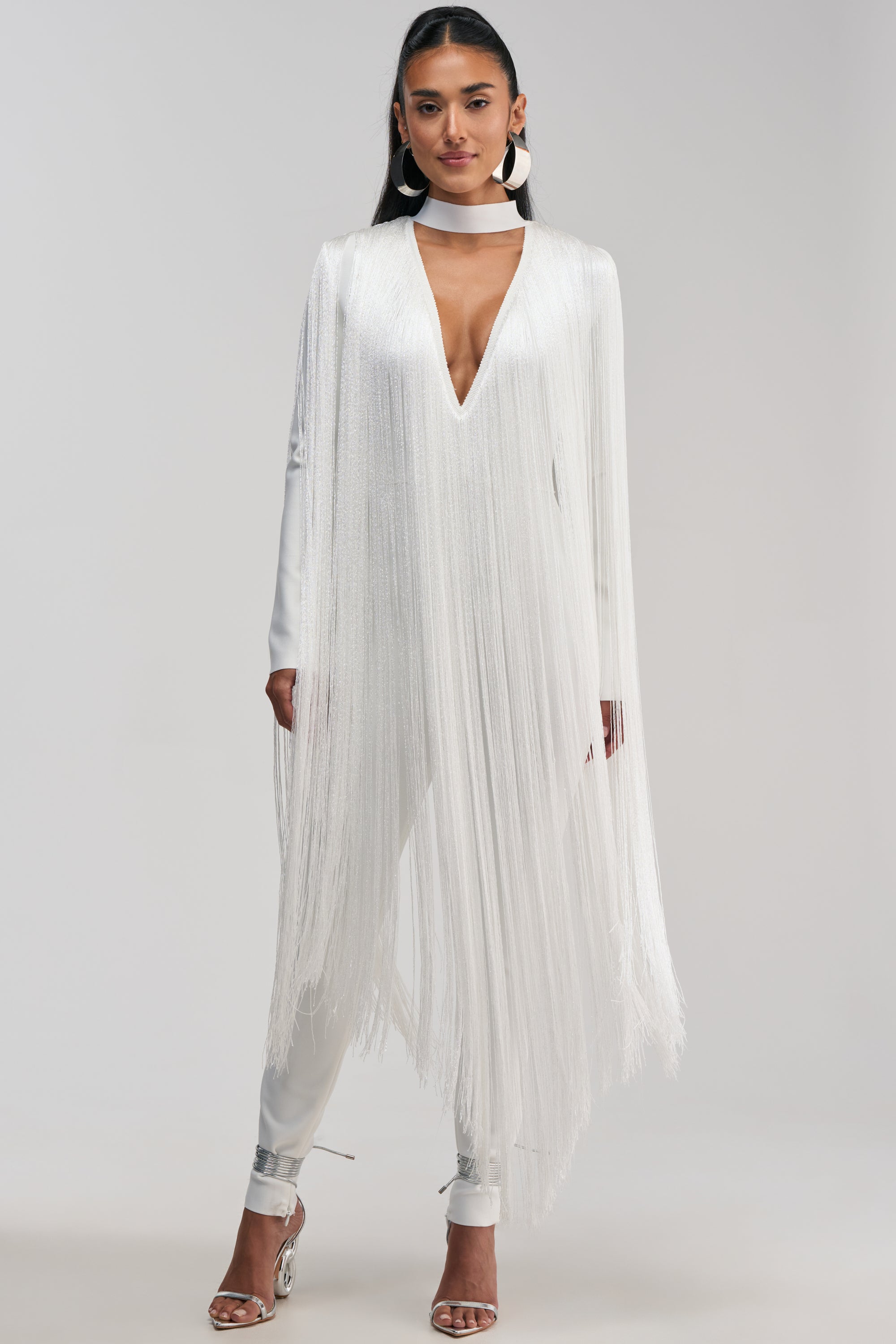 Dressed in the BELLE BANDAGE JUMPSUIT WITH FRINGE IN WHITE—a striking metallic one-piece featuring a deep V-neckline, long bandage-style sleeves, and bold fringe accents—a woman stands facing forward against a plain background. Paired with a matching choker and silver high heels, this eye-catching ensemble is the ultimate choice for a night out.