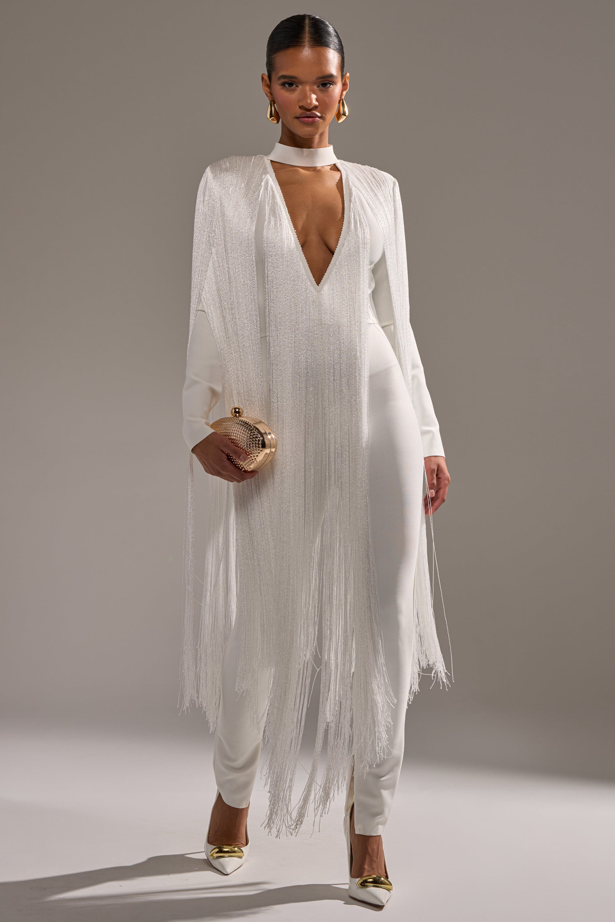 A woman stands confidently in the BELLE BANDAGE JUMPSUIT WITH FRINGE IN WHITE, featuring a deep V neckline and long, fringed cape detail. She accessorizes with a gold clutch and matching pointed heels against a plain gray background.