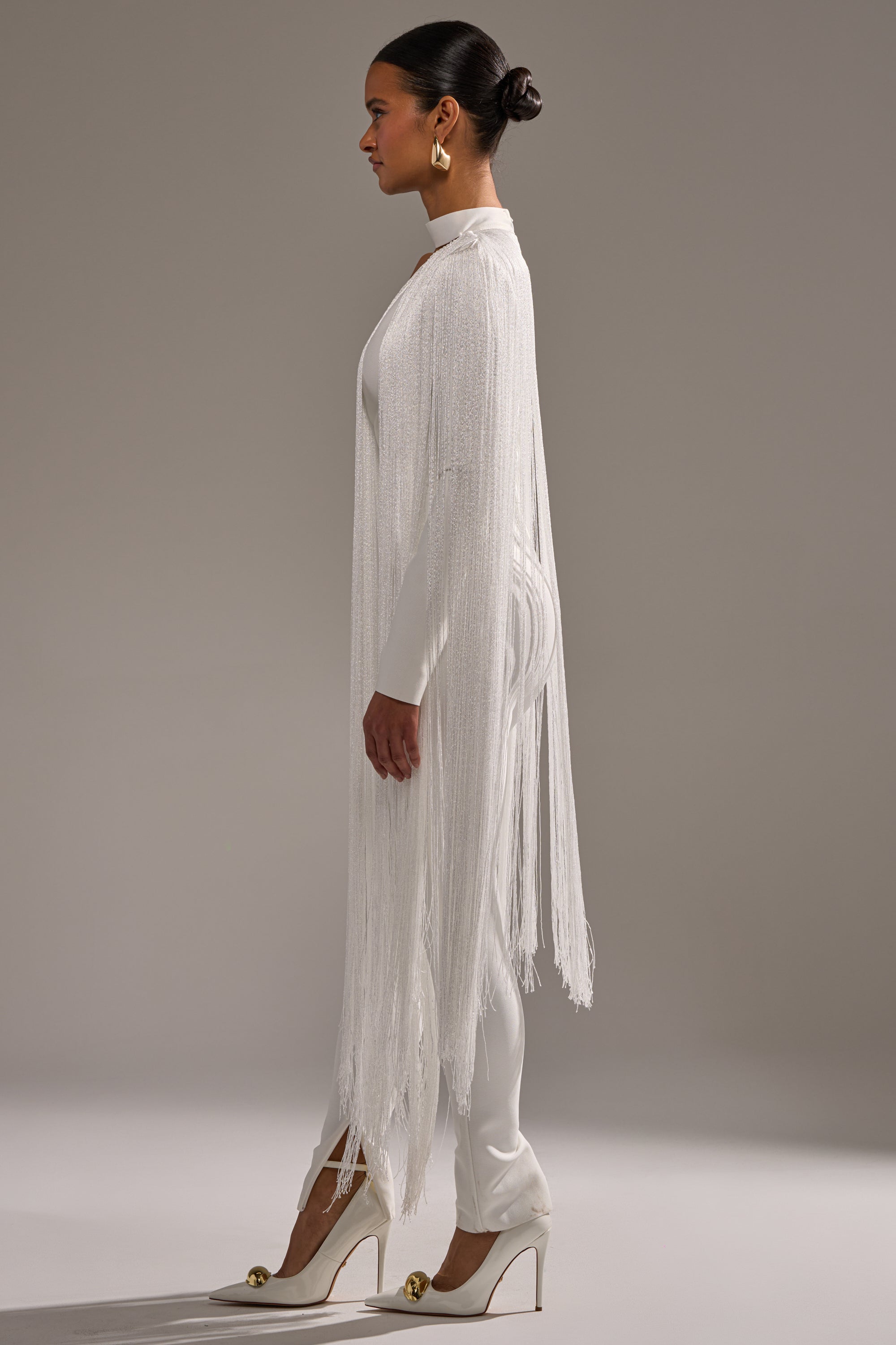 A woman in profile stands against a neutral background, wearing the BELLE BANDAGE JUMPSUIT WITH FRINGE IN WHITE. Her hair is in a sleek bun, styled with gold earrings and white heels featuring gold embellishments.
