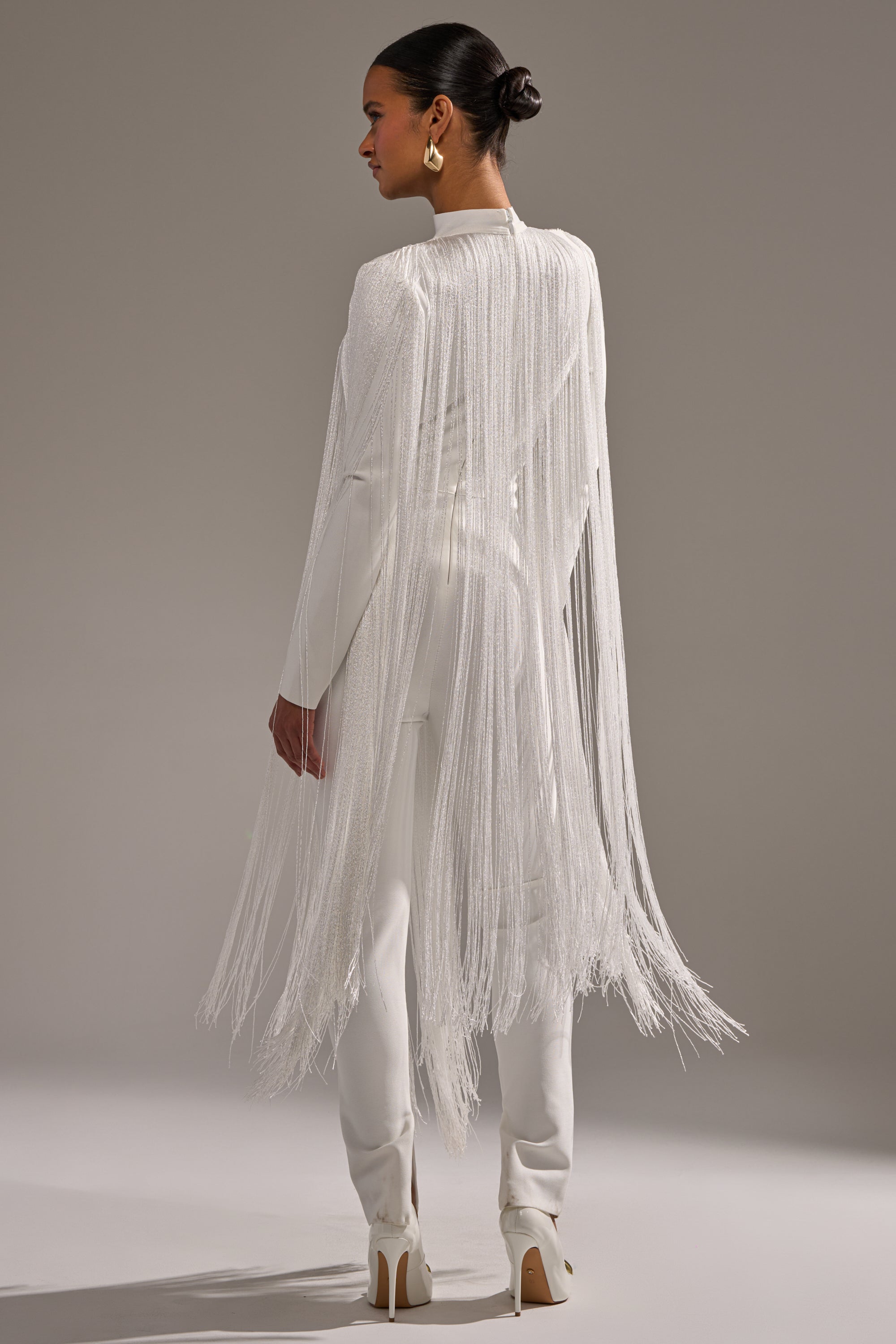 A woman faces away, wearing the BELLE BANDAGE JUMPSUIT WITH FRINGE IN WHITE. The chic white jumpsuit features long fringe from shoulders to below the knees, styled with a neat bun, statement gold earrings, and white heels.