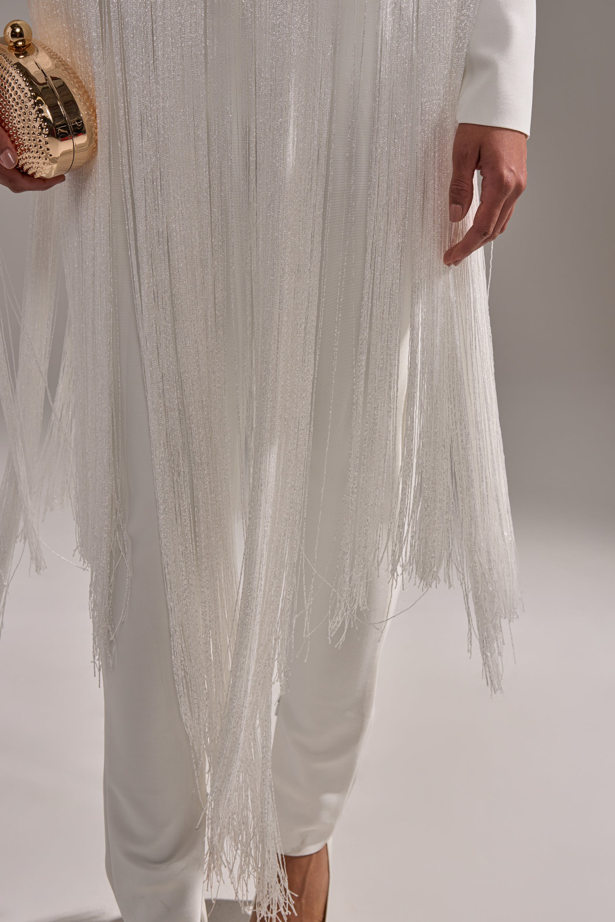 A person wearing the BELLE BANDAGE JUMPSUIT WITH FRINGE IN WHITE holds a gold clutch. Only the lower half of their body is visible against a neutral background.