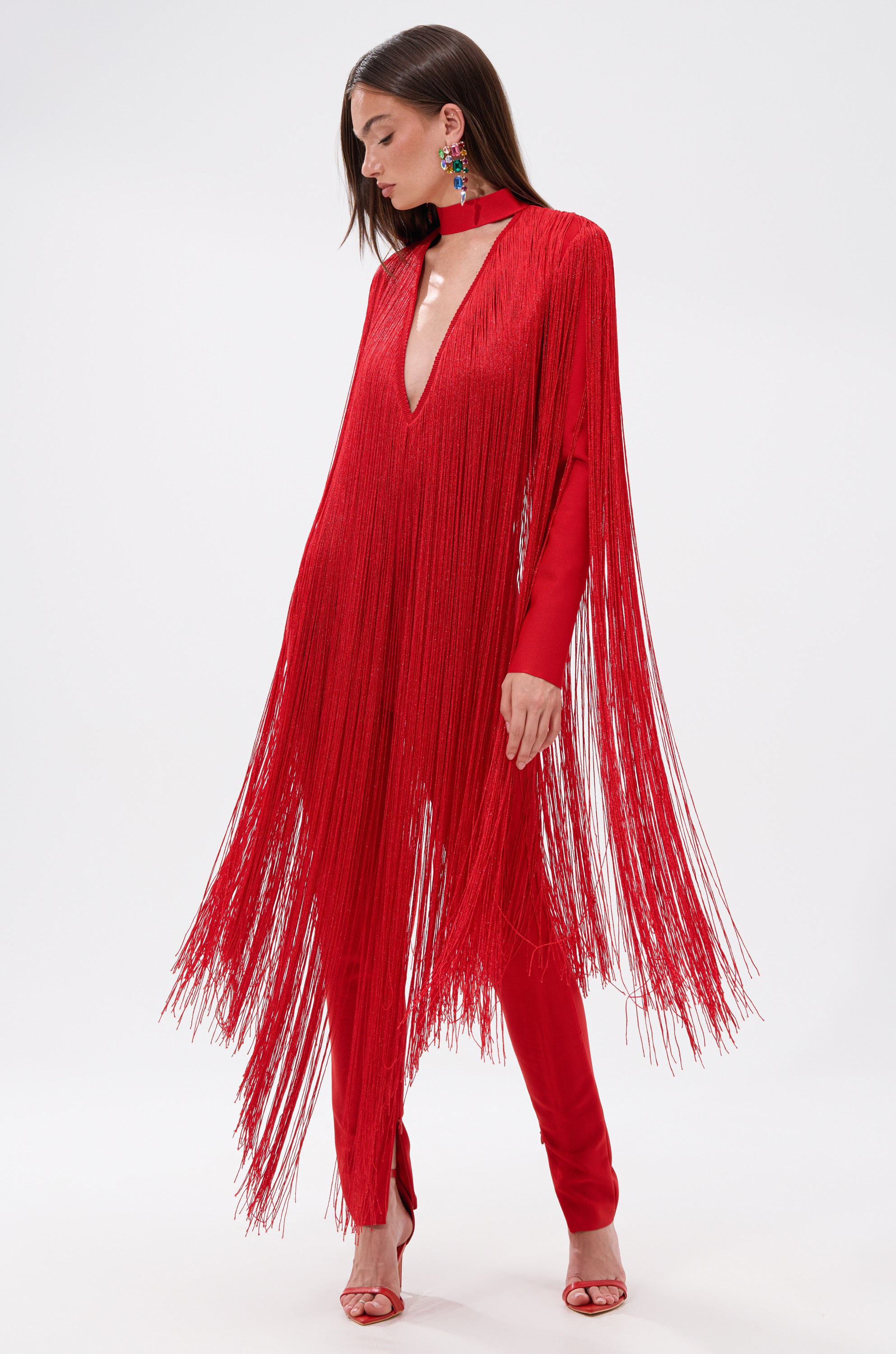 A woman wears the BELLE BANDAGE JUMPSUIT WITH FRINGE, featuring a vibrant red hue, deep V-neckline, and long flowing fringe. She pairs it with matching heels and colorful earrings, her long brown hair draped over one shoulder.