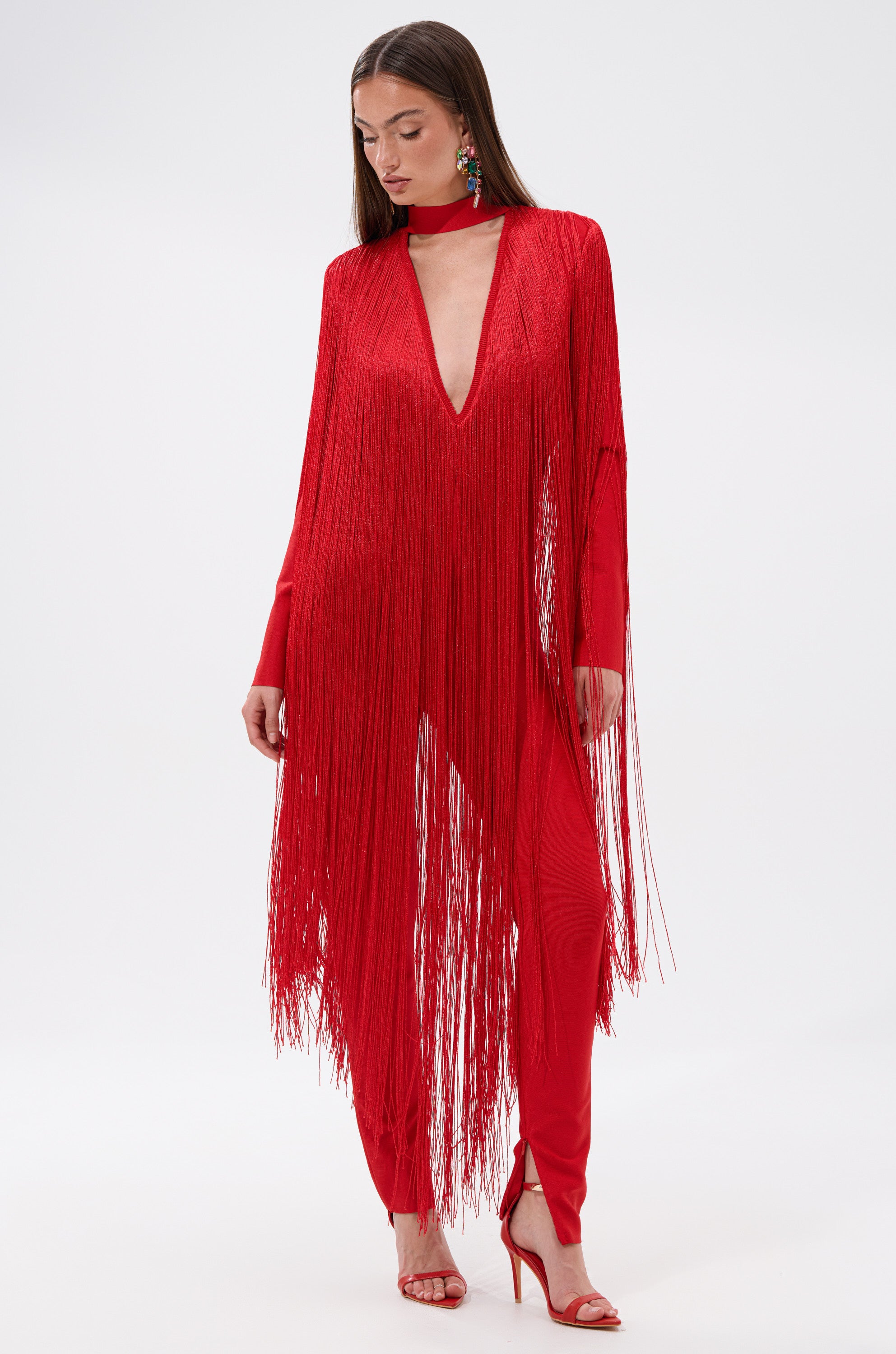 A woman poses against a white background wearing the BELLE BANDAGE JUMPSUIT WITH FRINGE, featuring a vibrant red hue, deep V-neckline, fitted pants, and paired with red heels and statement earrings.