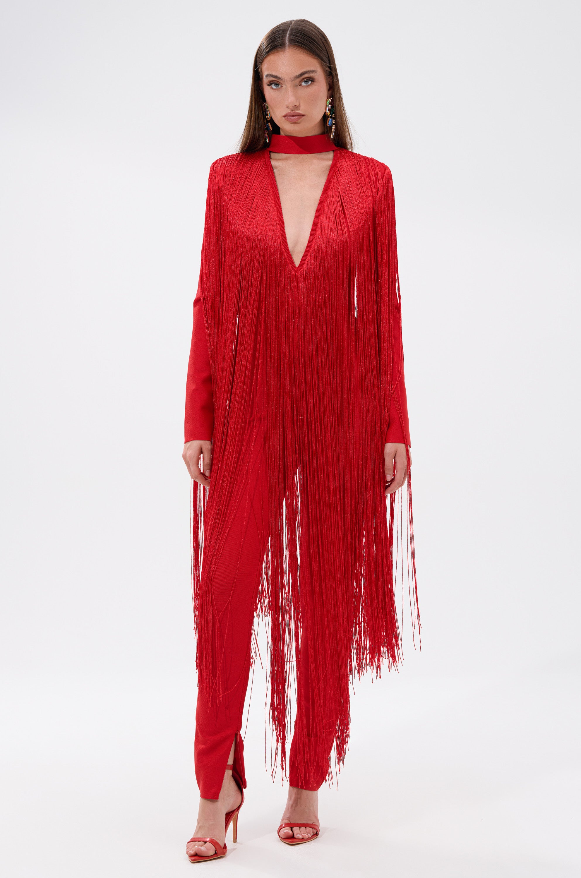 A woman poses confidently against a plain white background in the BELLE BANDAGE JUMPSUIT WITH FRINGE, styled in bold red with a deep V neckline and matching red high-heeled sandals.