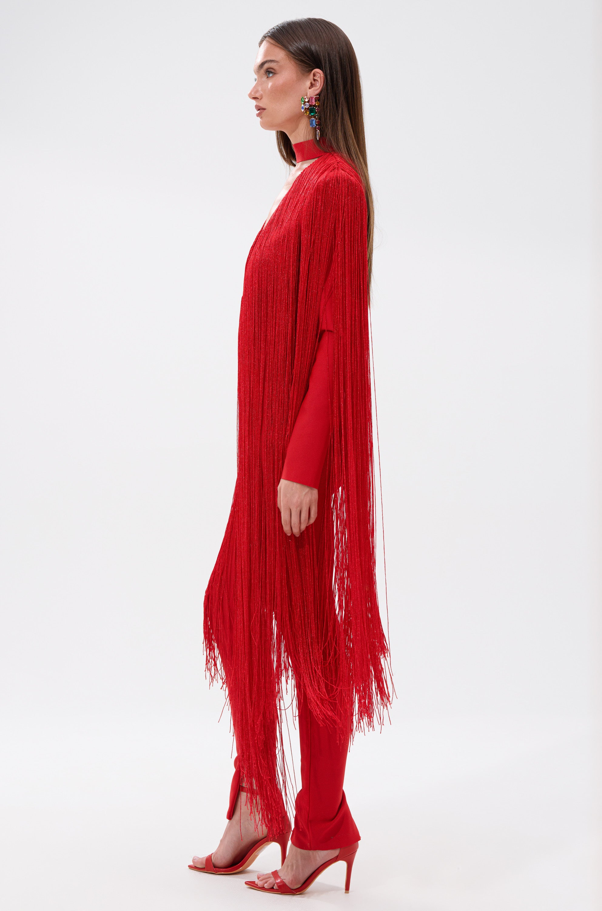 A woman stands in profile against a white background wearing the BELLE BANDAGE JUMPSUIT WITH FRINGE in bright red, paired with matching heels. Her straight brown hair is down, and she accessorizes with colorful statement earrings.