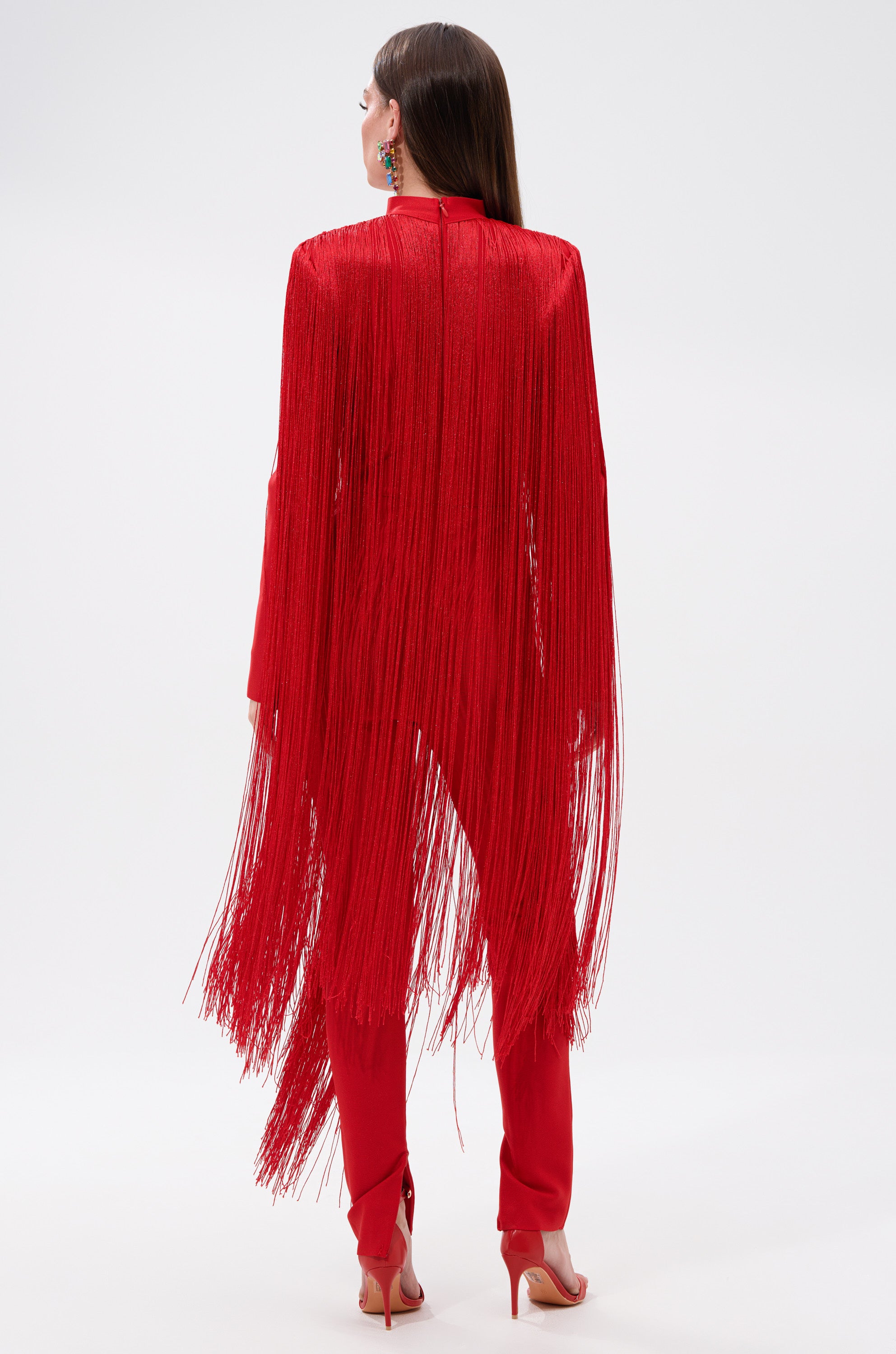 A woman with straight brown hair and high heels stands against a white background, wearing the BELLE BANDAGE JUMPSUIT WITH FRINGE—a vibrant red jumpsuit featuring long, flowing fringe draping from shoulders to legs.