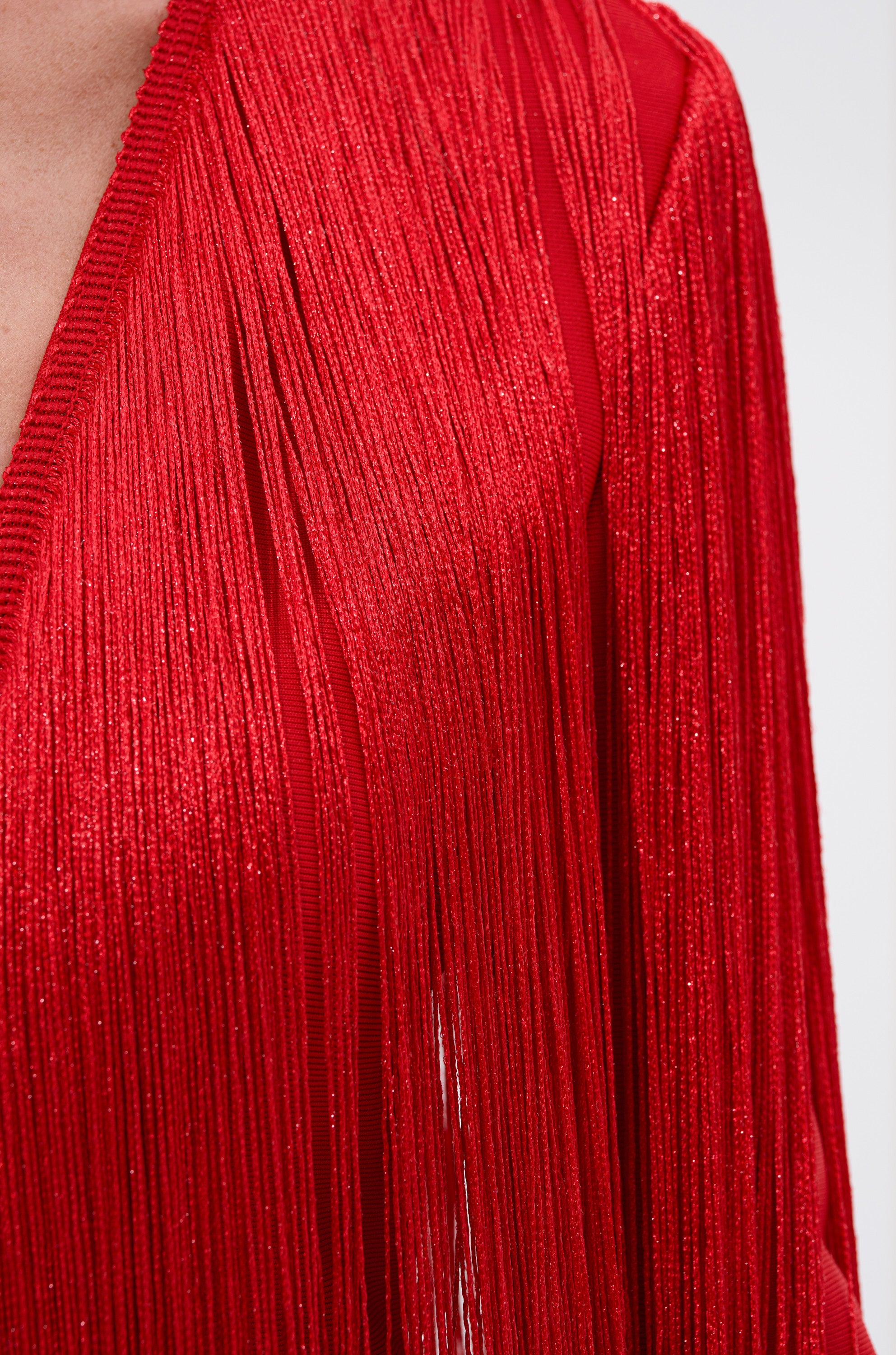 Close-up of a person wearing the BELLE BANDAGE JUMPSUIT WITH FRINGE, featuring vibrant red fabric and long, shiny fringe details that create a textured and dynamic look. Only the shoulder and torso are visible.
