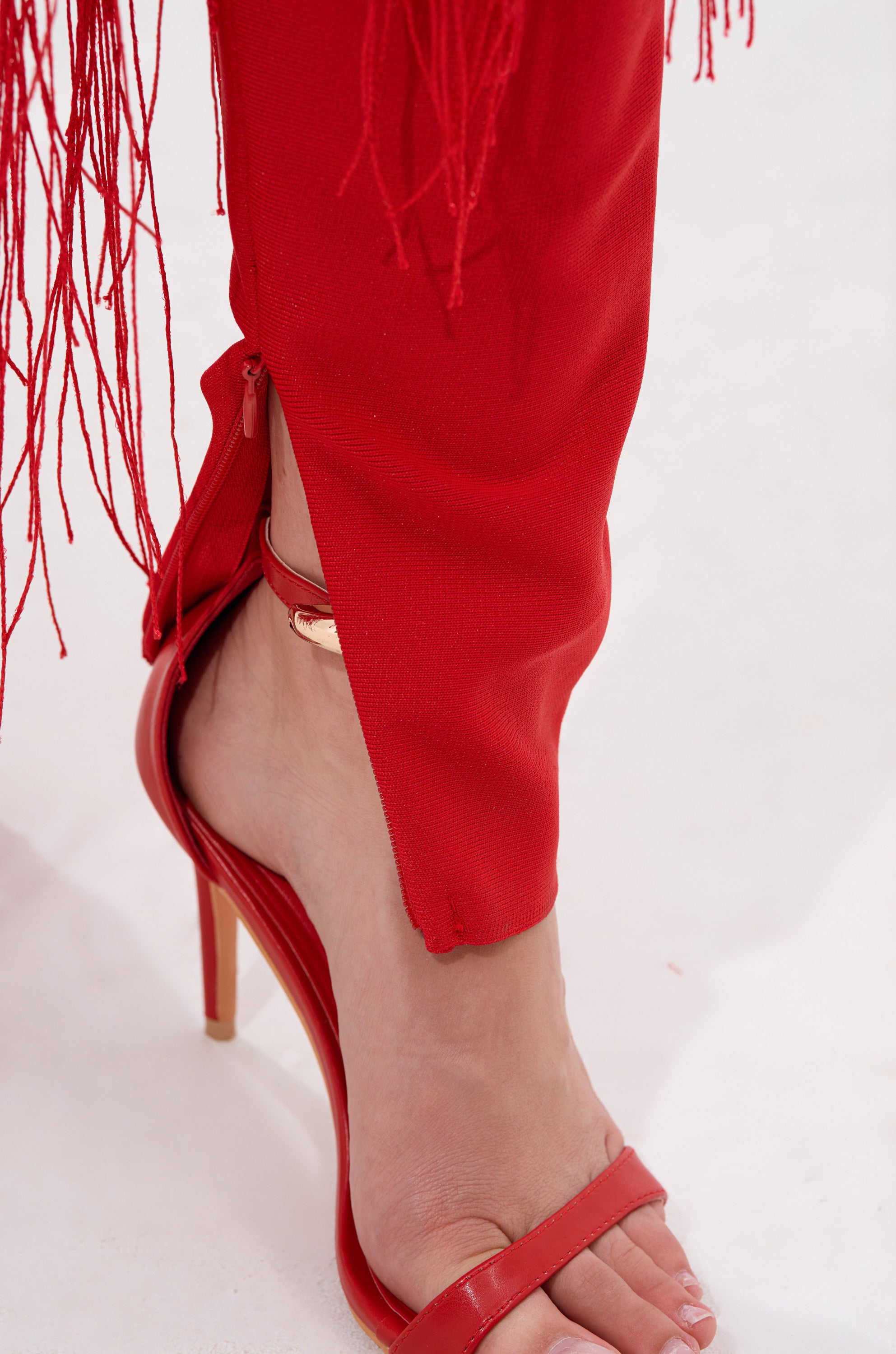 A close-up of a foot in a red high-heeled sandal and BELLE BANDAGE JUMPSUIT WITH FRINGE pants with an ankle zipper, set against a white background.