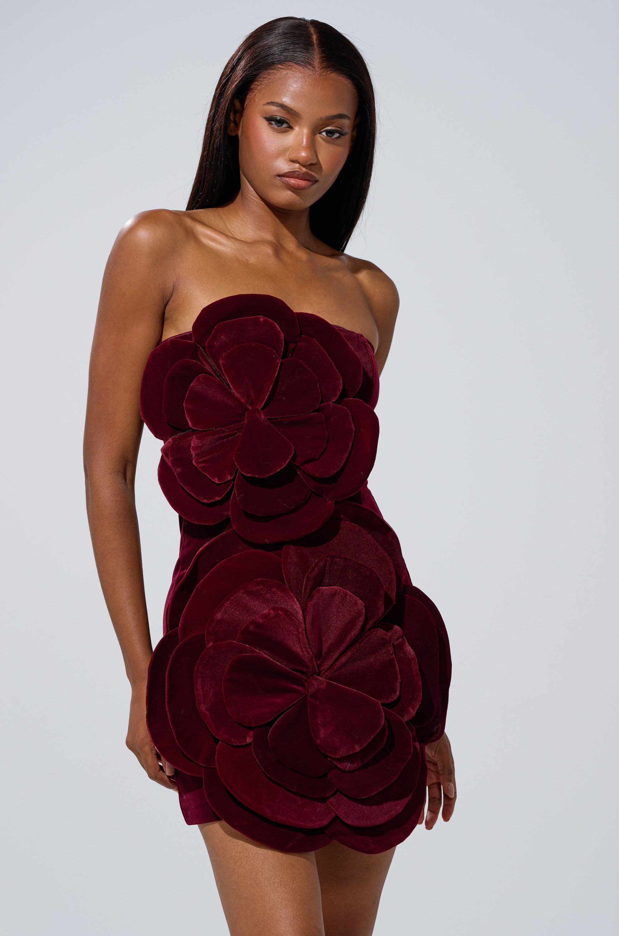 A woman stands against a plain gray background, confidently wearing the FLOWER POWER VELVET MINI DRESS—a strapless mini dress with large, layered floral appliqué. Her long, straight hair complements the bold look.