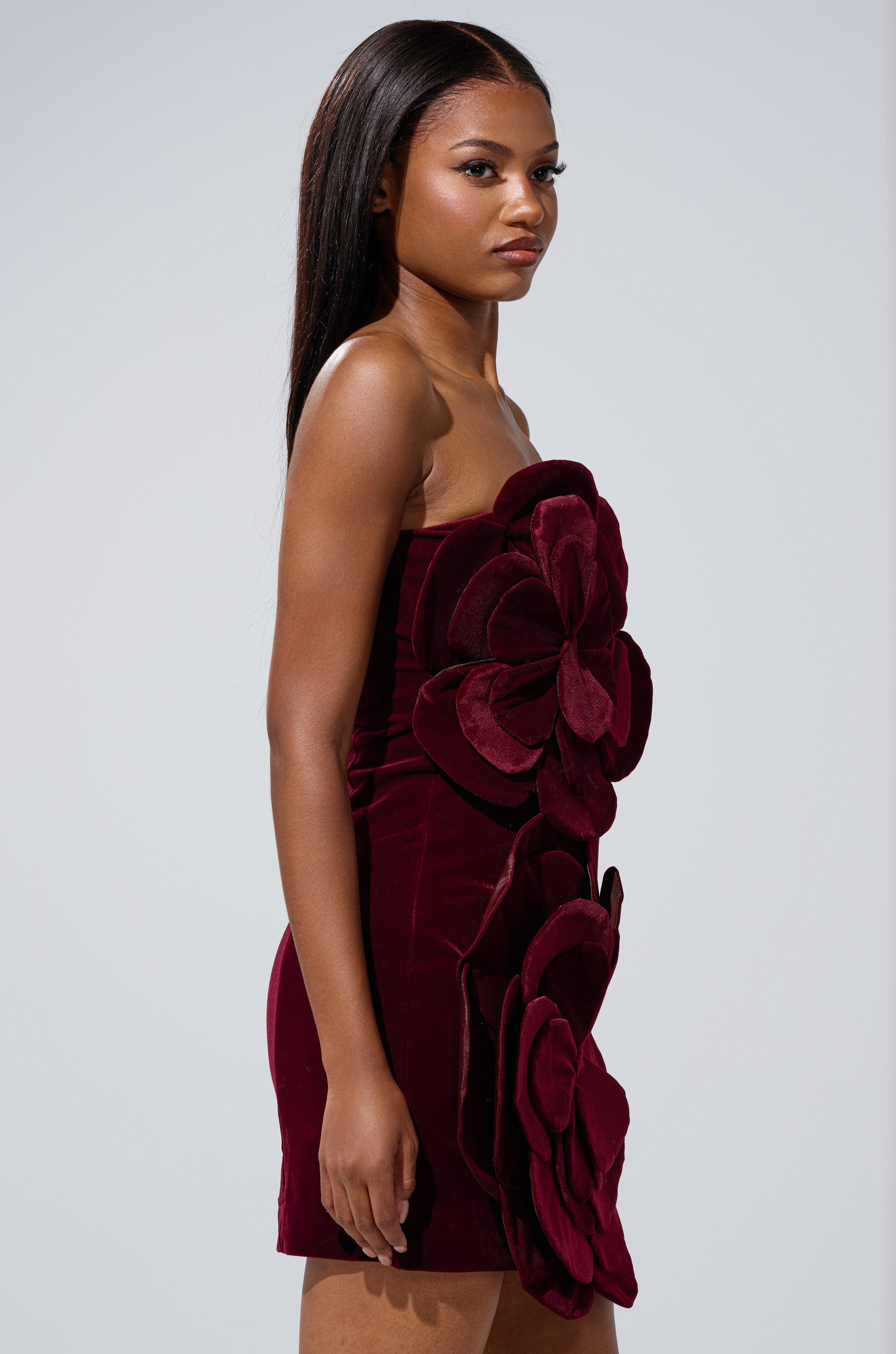 A woman with long straight hair and a neutral expression stands sideways against a plain background, modeling the FLOWER POWER VELVET MINI DRESS featuring a strapless neckline and bold 3D floral appliqués on the front.