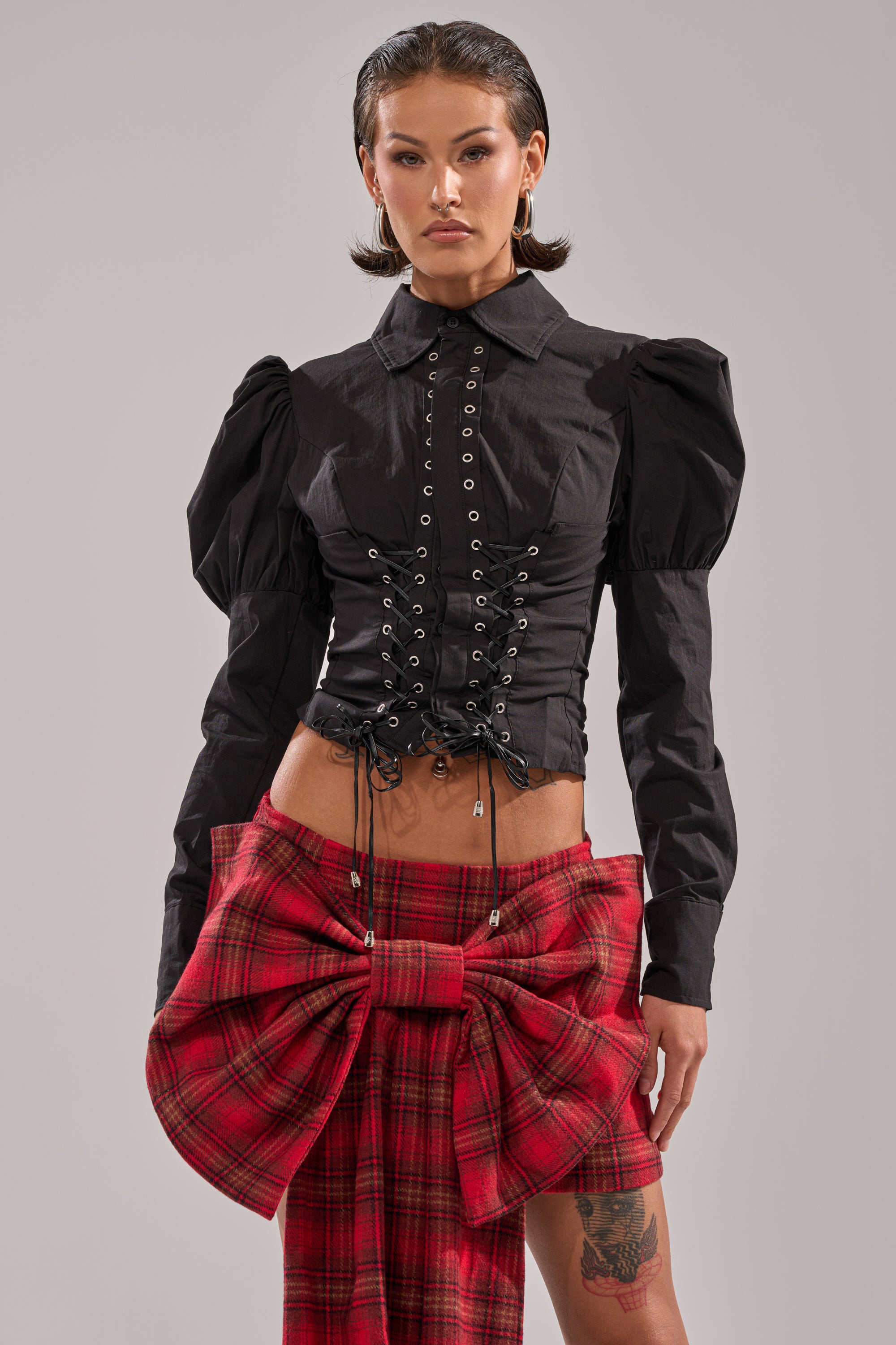 A woman with slicked-back hair wears the DAKOTA LACE UP BUTTON-DOWN BLOUSE IN BLACK, styled with puffed sleeves and a lace-up front, paired with a red plaid skirt featuring a large bow at the waist. She stands facing forward against a plain backdrop.