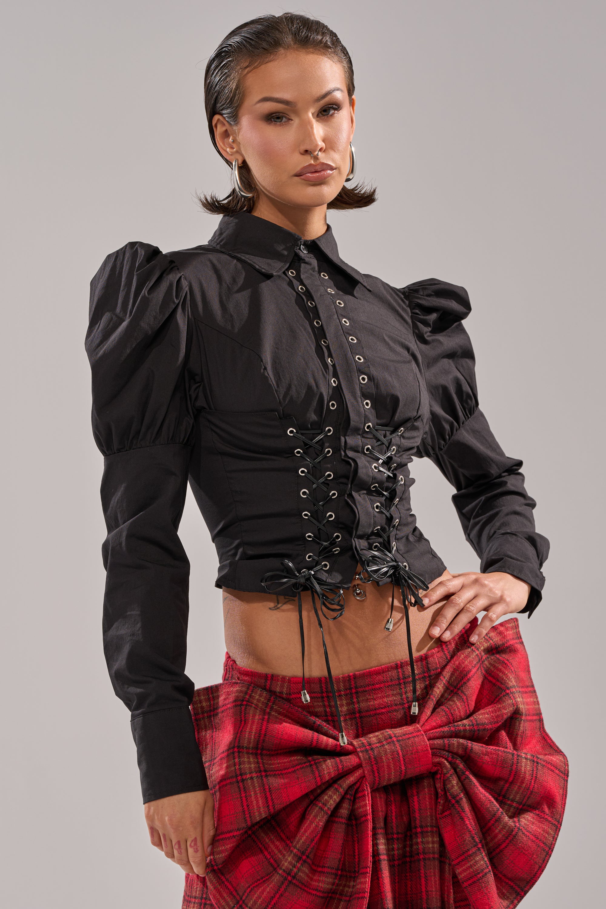 A woman poses confidently in the DAKOTA LACE UP BUTTON-DOWN BLOUSE IN BLACK, styled with a red plaid skirt with a large bow. She wears her hair sleek and slicked-back, finishing the look with hoop earrings.