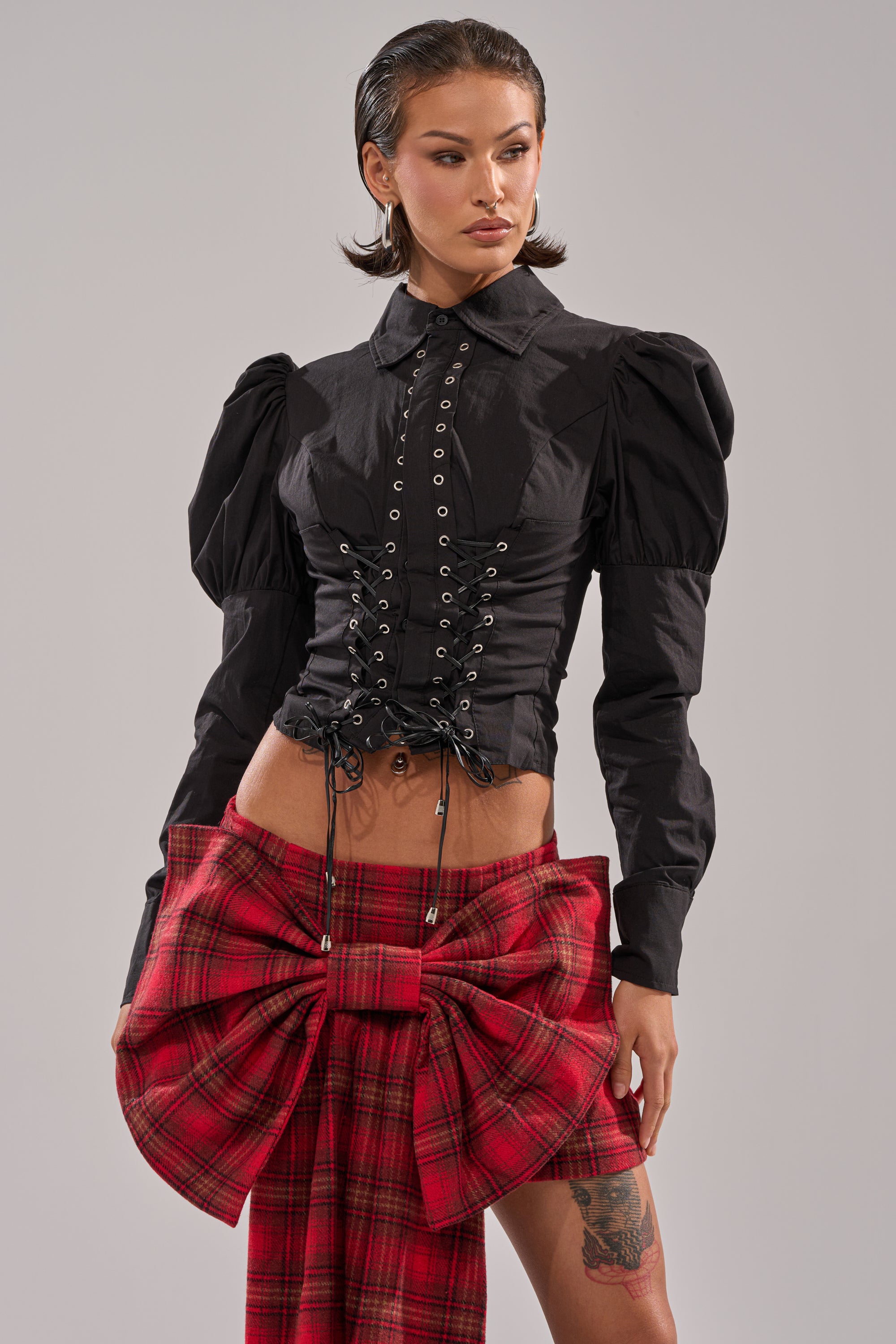 A woman wears the DAKOTA LACE UP BUTTON-DOWN BLOUSE IN BLACK, styled with a red plaid skirt with an oversized front bow, standing against a plain light background.