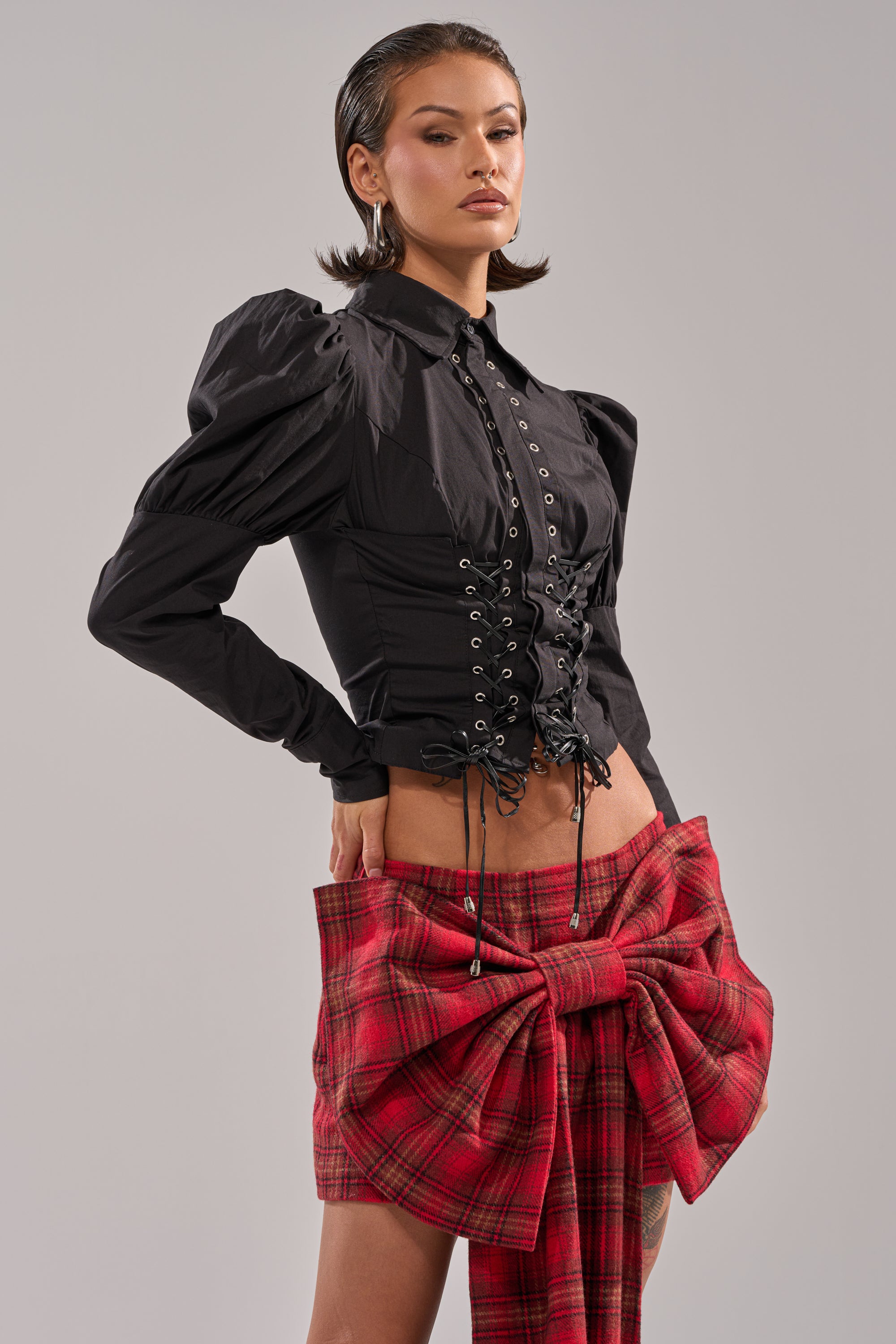 A woman poses confidently in the DAKOTA LACE UP BUTTON-DOWN BLOUSE IN BLACK, styled with a red plaid skirt with a dramatic waist bow, against a plain light gray background.