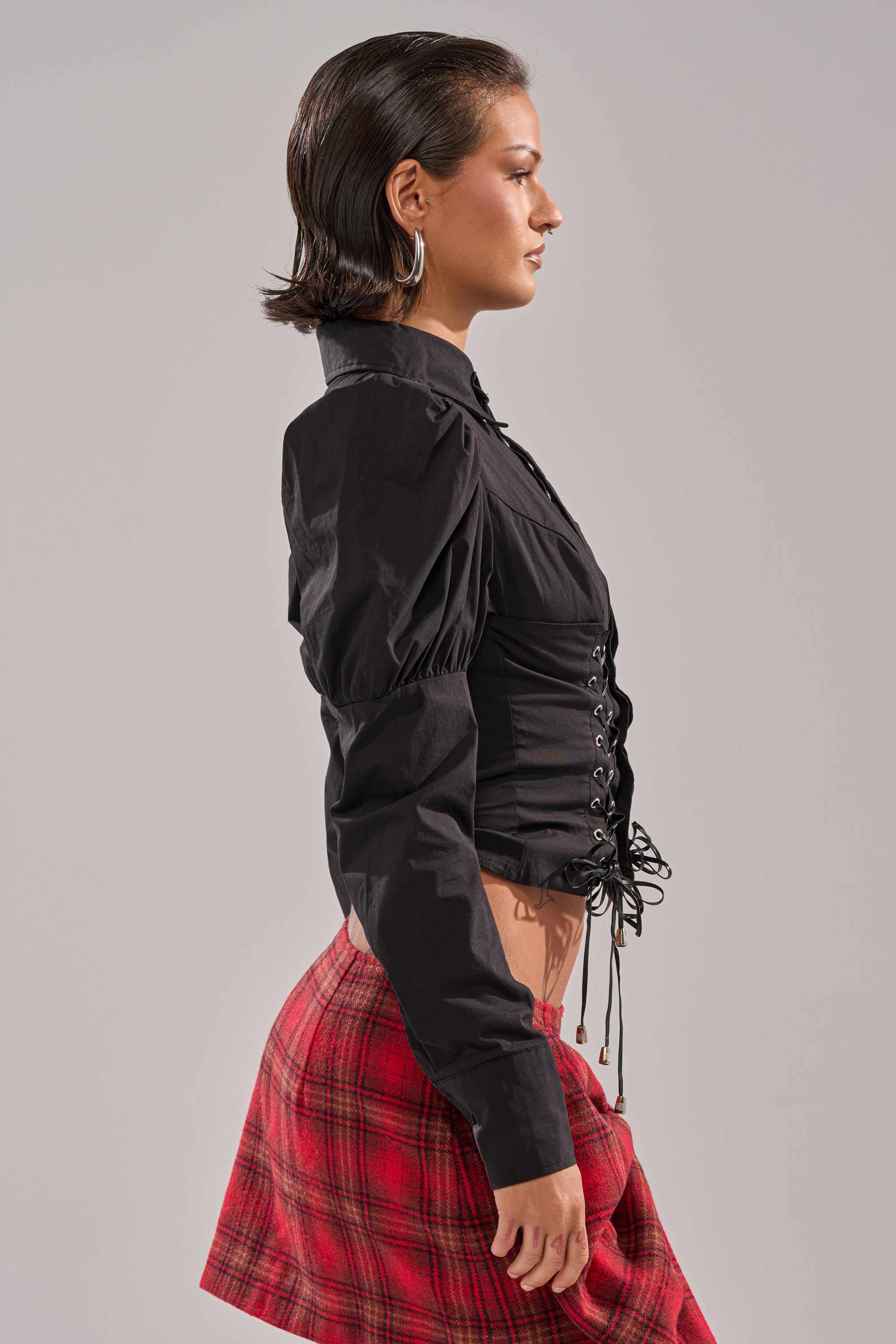 A woman in profile wears the DAKOTA LACE UP BUTTON-DOWN BLOUSE IN BLACK with puffed shoulders and lace-up detail, paired with a red plaid mini skirt. She has short slicked-back hair and large hoop earrings, standing against a plain background.