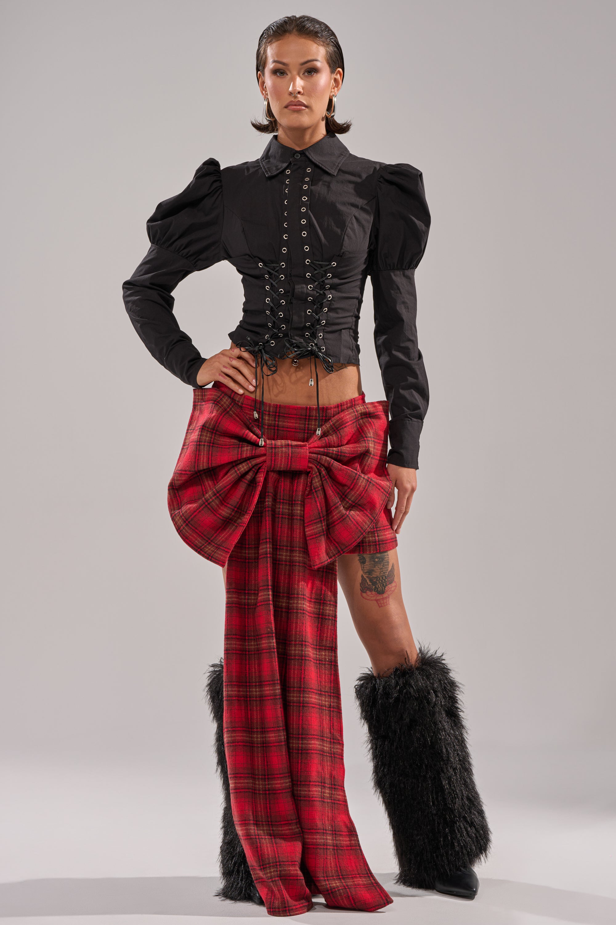 A model confidently wears the DAKOTA LACE UP BUTTON-DOWN BLOUSE IN BLACK, styled with red plaid pants featuring a large waist bow and black furry leg warmers, standing against a plain light gray background.