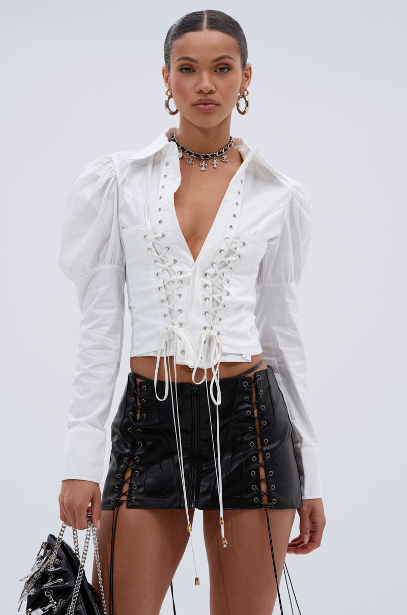 A woman wears the DAKOTA LACEUP BUTTON DOWN BLOUSE styled with a black mini skirt featuring lace-up details. She accessorizes with hoop earrings, layered necklaces, and carries a chic handbag.
