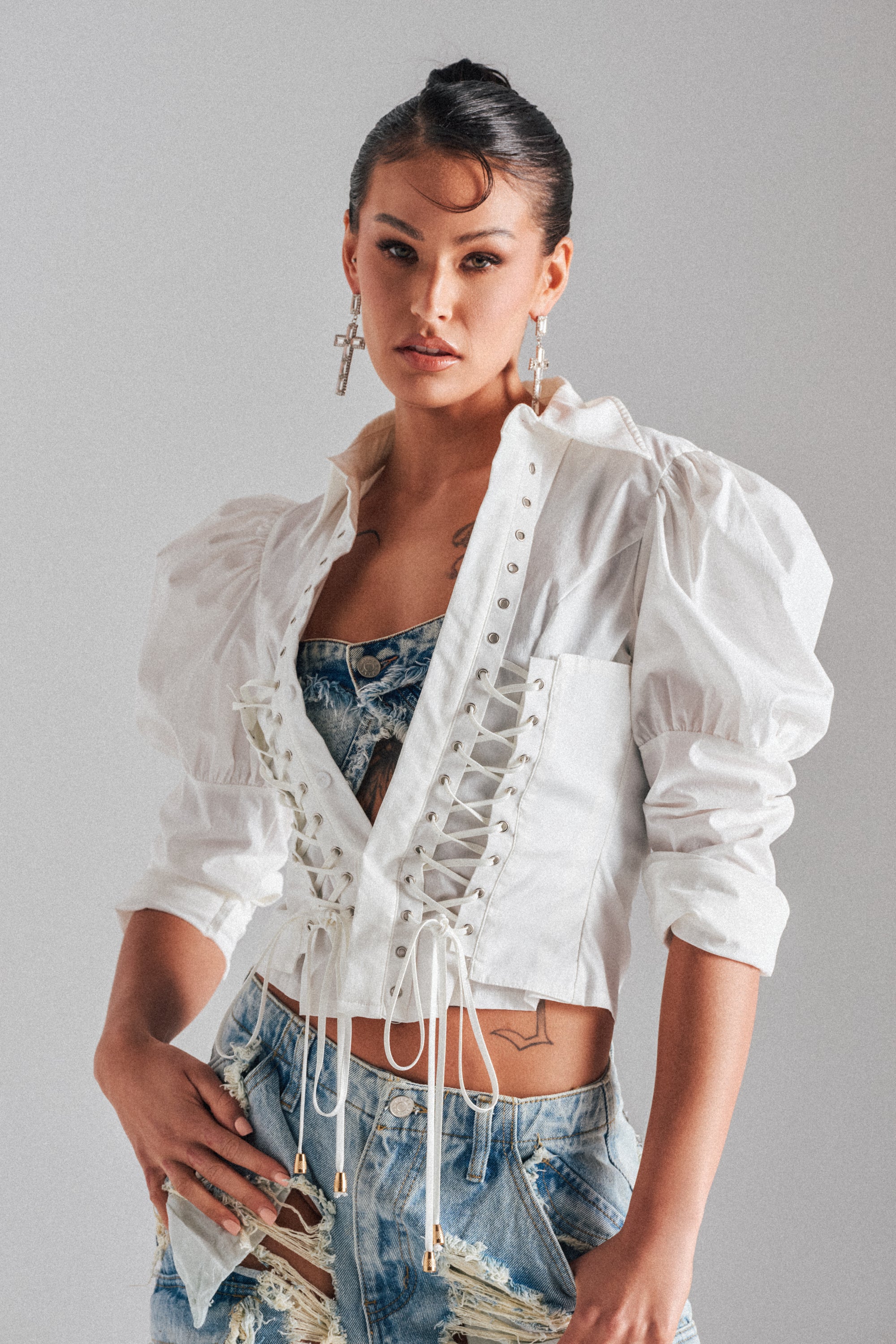 A woman poses in the DAKOTA LACEUP BUTTON DOWN BLOUSE over a denim top and distressed shorts, accessorized with hoop and cross earrings. Her hair is styled up with a wave in front, set against a plain light background.