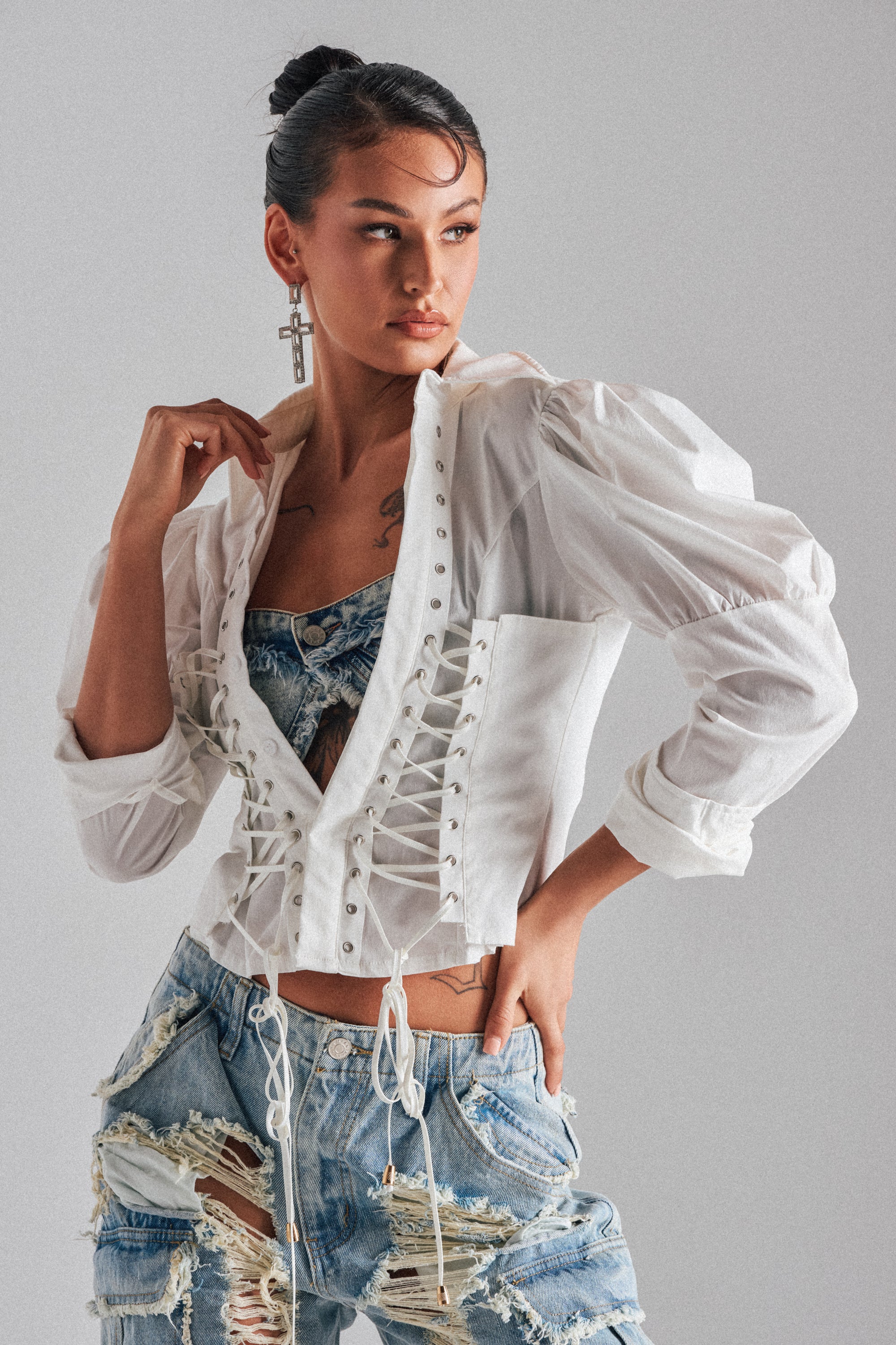A woman poses confidently in the DAKOTA LACEUP BUTTON DOWN BLOUSE over a patterned top and distressed jeans. Her hair is in a bun, she wears large cross earrings, and stands against a plain light gray background.