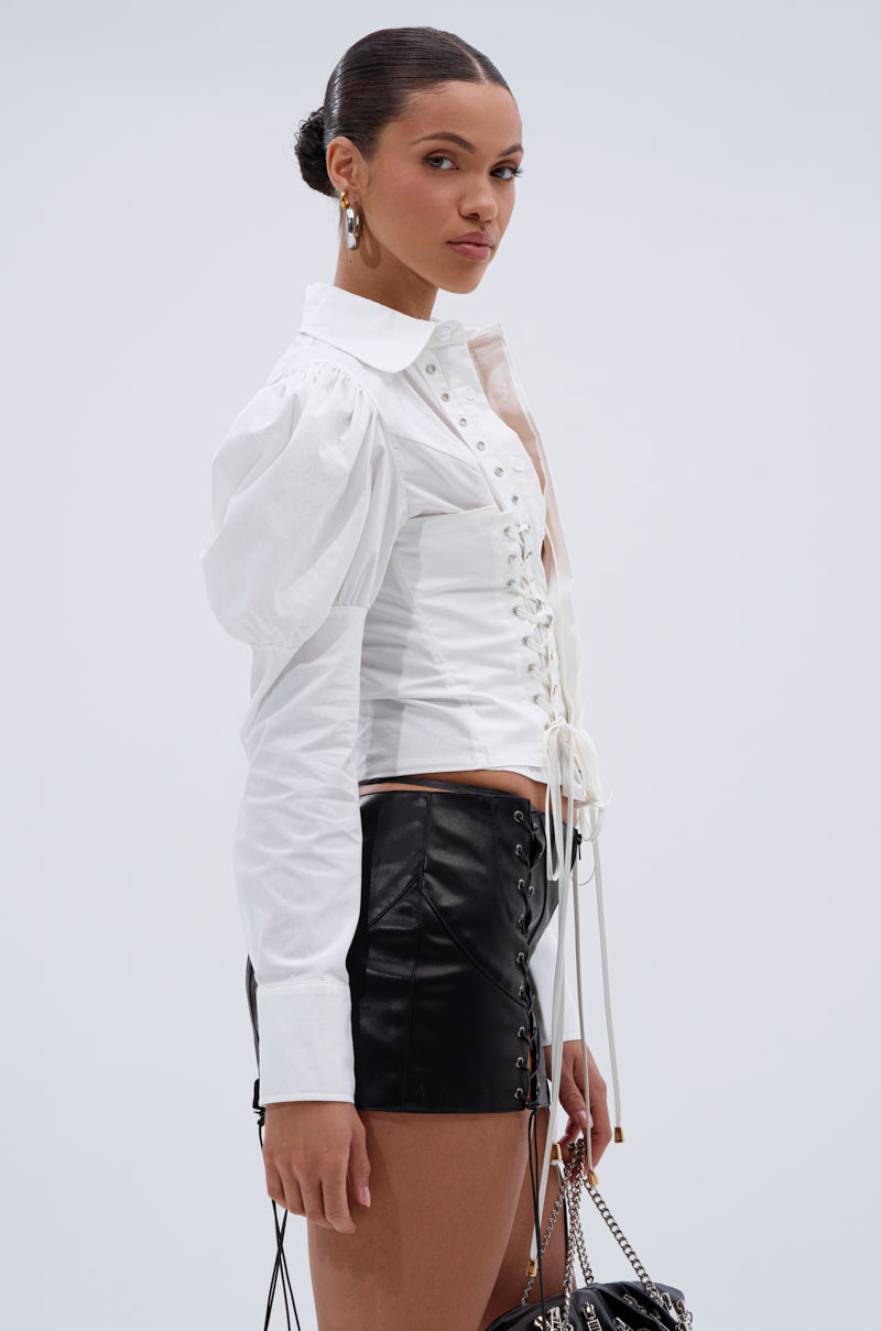 A woman, hair in a sleek bun, models the DAKOTA LACEUP BUTTON DOWN BLOUSE—a white puff-sleeve, collared top with lace-up details—paired with black high-waisted leather shorts and a chain-strap bag.