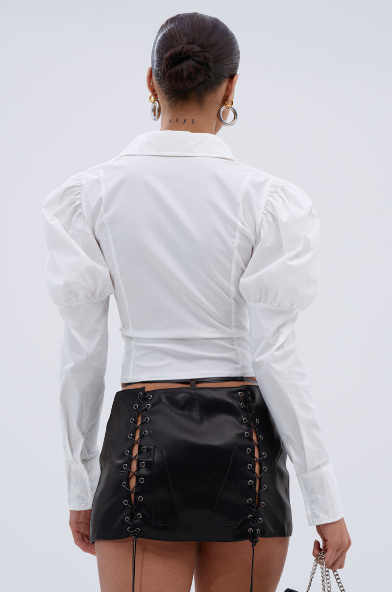 A person with hair in a sleek bun wears the DAKOTA LACEUP BUTTON DOWN BLOUSE and a black mini skirt. They accessorize with hoop earrings, show off a neck tattoo, and carry a chain-strap purse.