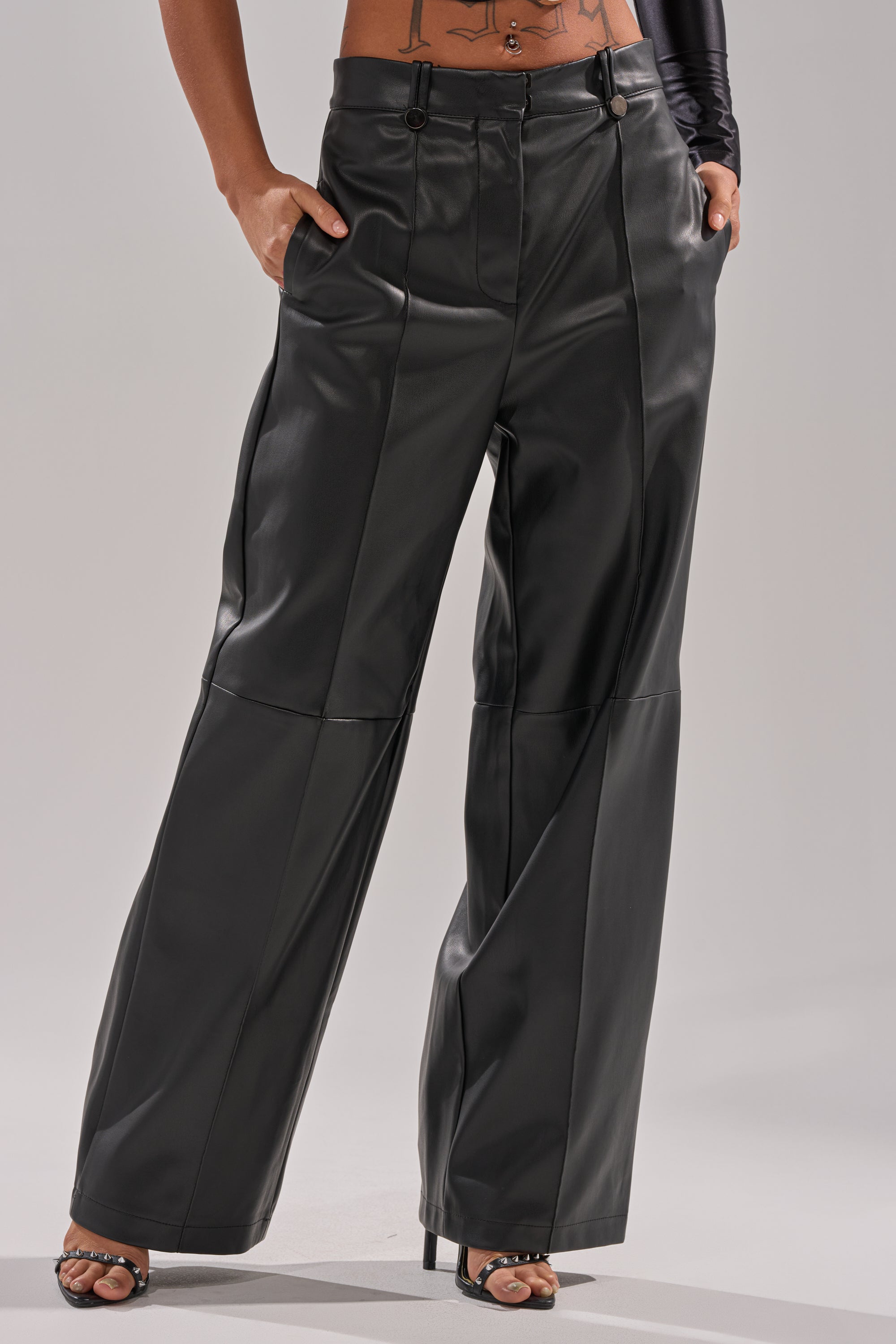 A person styles the FAUX STRETCHY LEATHER TROUSER—high-waisted, black, with seam details and a straight-leg fit—with black open-toe heels and a cropped top, hands in pockets, standing against a plain background.