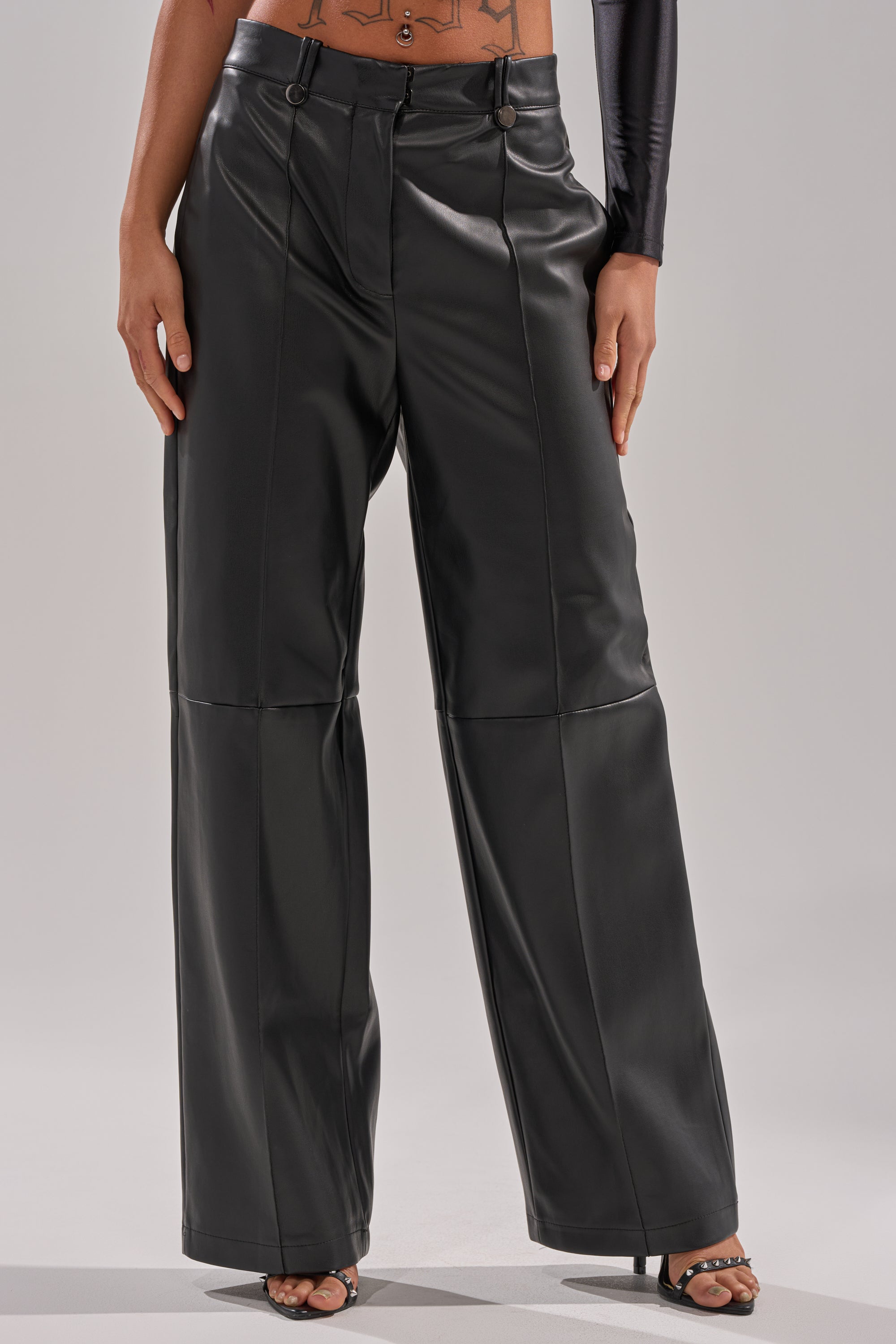 A person wears the FAUX STRETCHY LEATHER TROUSER—high-waisted, black, straight-leg faux leather—paired with heeled sandals, hands at sides, shown from shoulders to feet against a plain background.