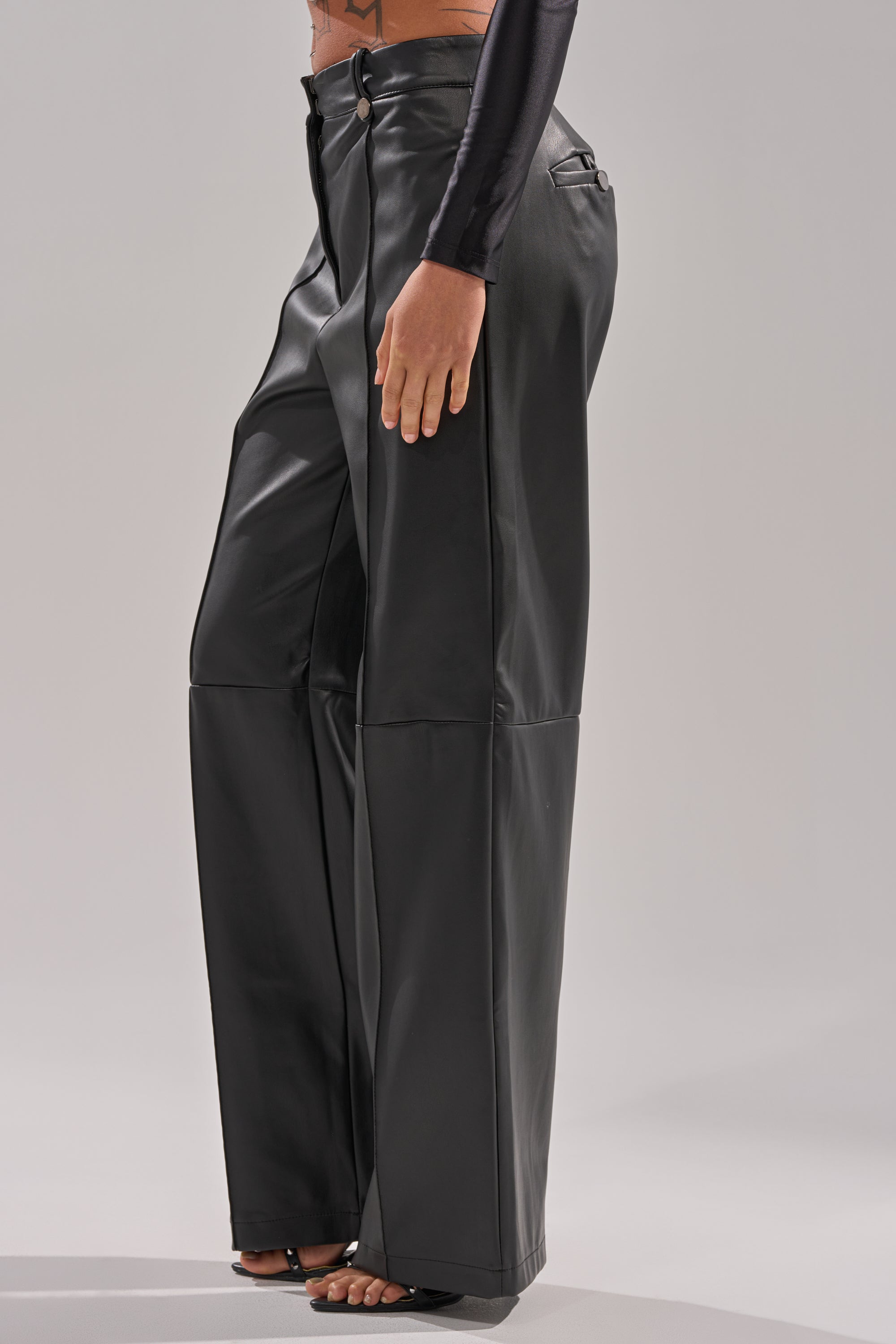 Someone, visible from the waist down, models the FAUX STRETCHY LEATHER TROUSER in black with a high-rise fit and wide legs, paired with open-toe heels against a plain light gray background.