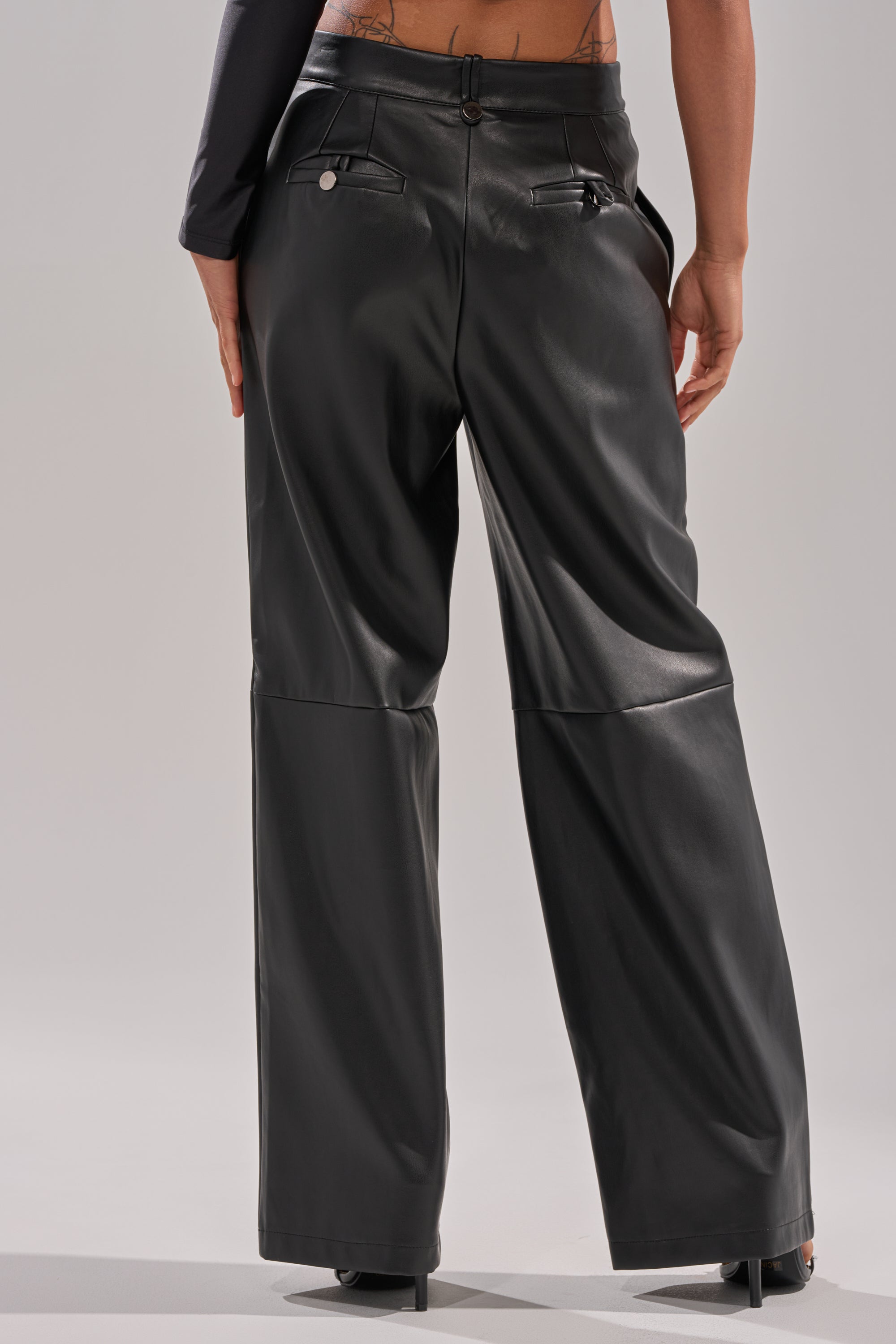A person is shown from behind wearing the FAUX STRETCHY LEATHER TROUSER in black, featuring a high-rise fit, straight-leg silhouette, shiny finish, back pockets, and paired with heeled shoes.