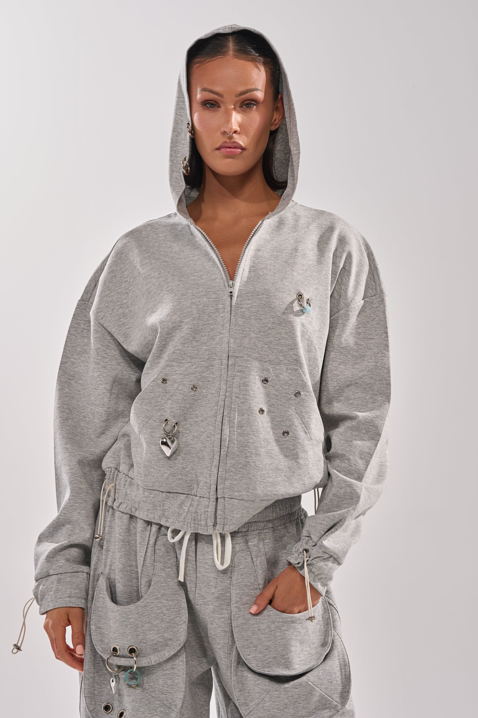 A woman stands facing forward in the GET WITH IT ZIP UP SWEATSHIRT, a light gray streetwear hoodie with the hood up, paired with matching cargo pants featuring multiple pockets and silver accents. She wears a neutral expression against a plain background. 0