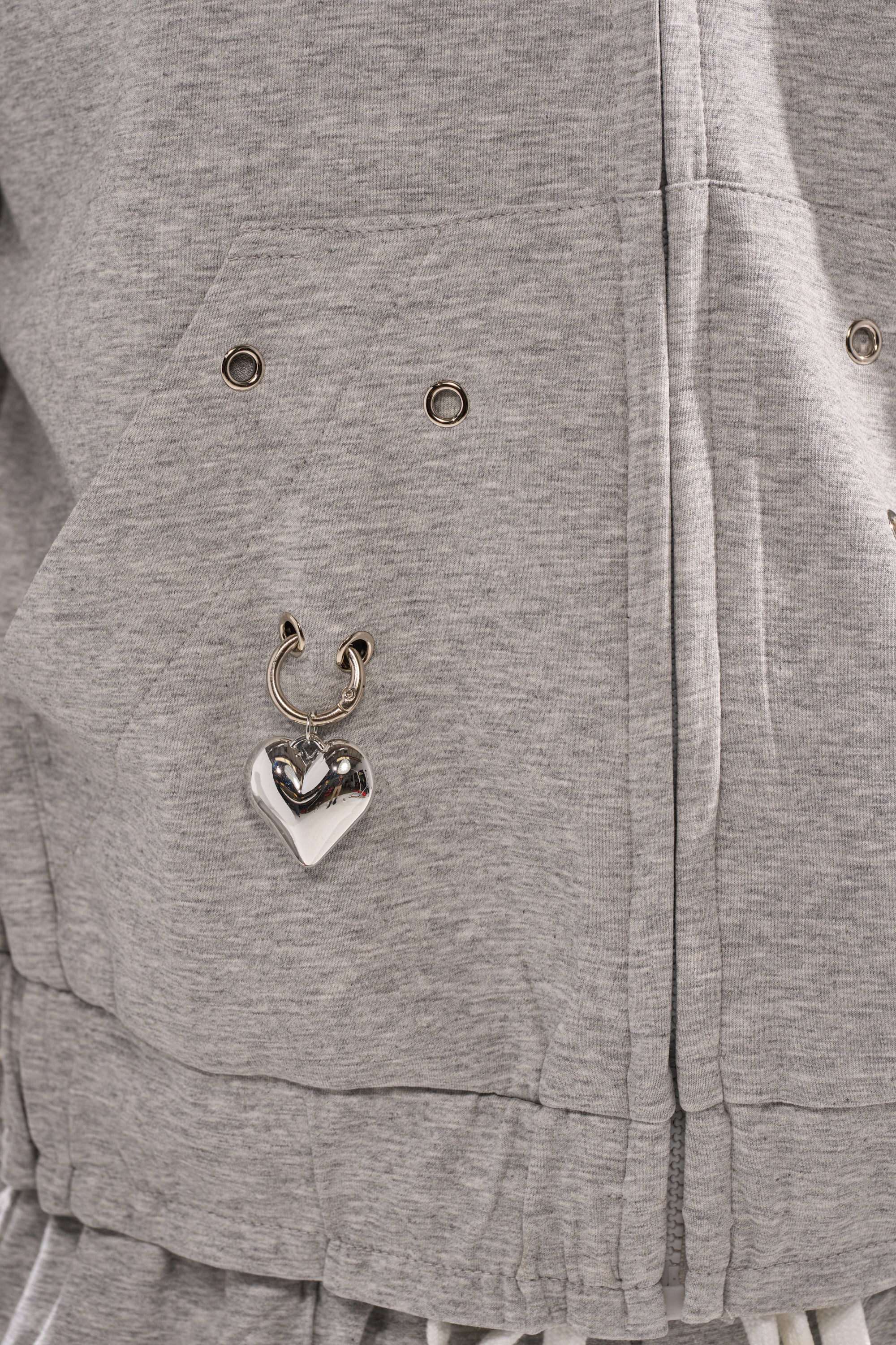 Close-up of the GET WITH IT ZIP UP SWEATSHIRT, a light gray streetwear hoodie featuring metal grommets and a shiny silver heart-shaped charm near the pocket. 10