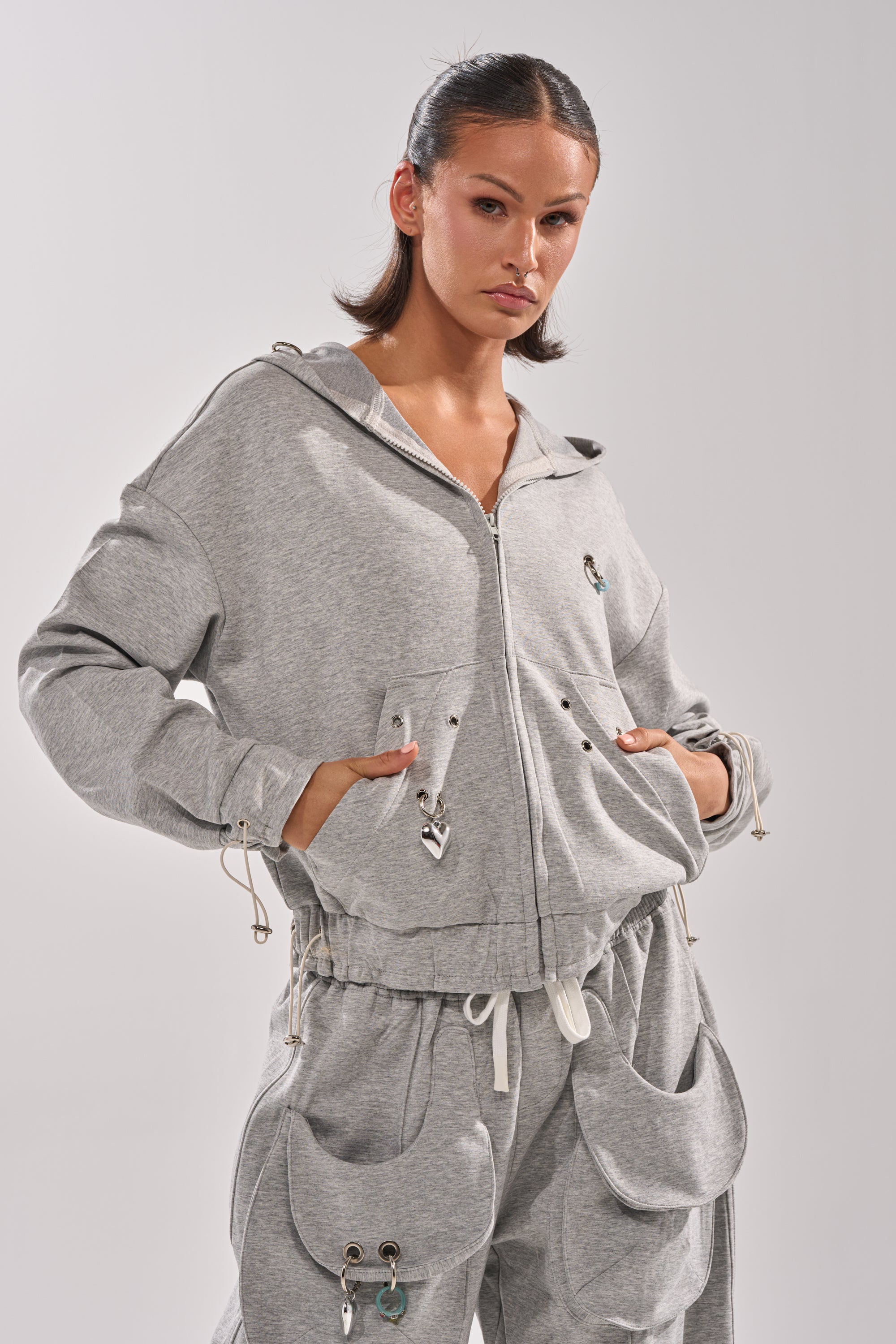 A woman stands confidently with her hands in the pockets of the GET WITH IT ZIP UP SWEATSHIRT, a light gray fleece hoodie featuring drawstrings and large front pockets, paired with matching sweatpants against a plain background. 3