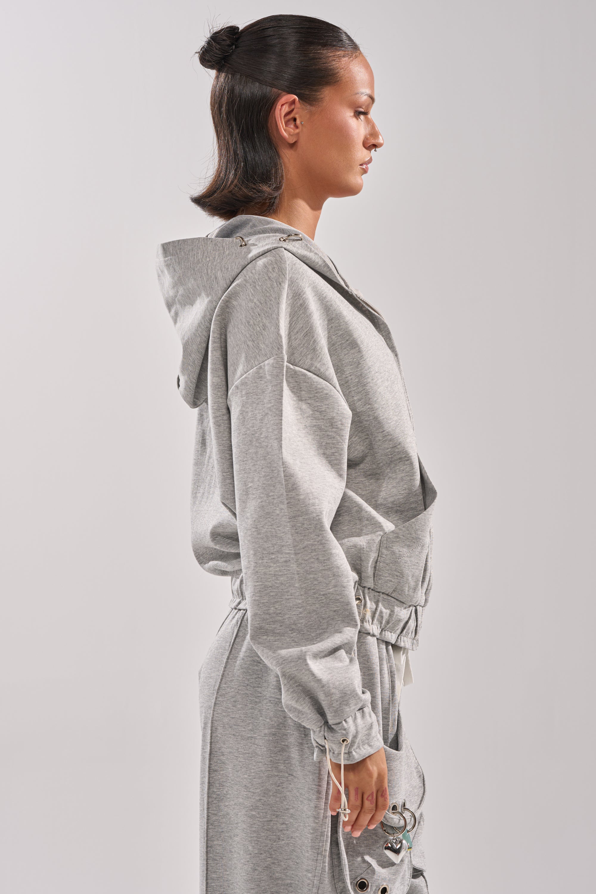 A woman with dark hair in a bun, wearing the GET WITH IT ZIP UP SWEATSHIRT in light gray, stands in profile against a plain light background. 4