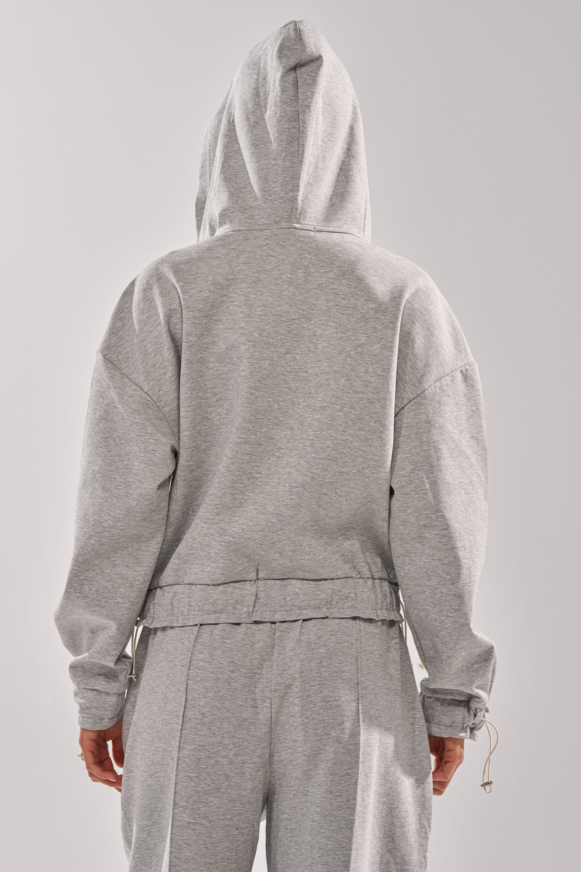 A person is shown from the back wearing the light gray GET WITH IT ZIP UP SWEATSHIRT, hood up and hands relaxed at their sides. The matching sweatpants complete the streetwear look against a plain, light-colored background. 6
