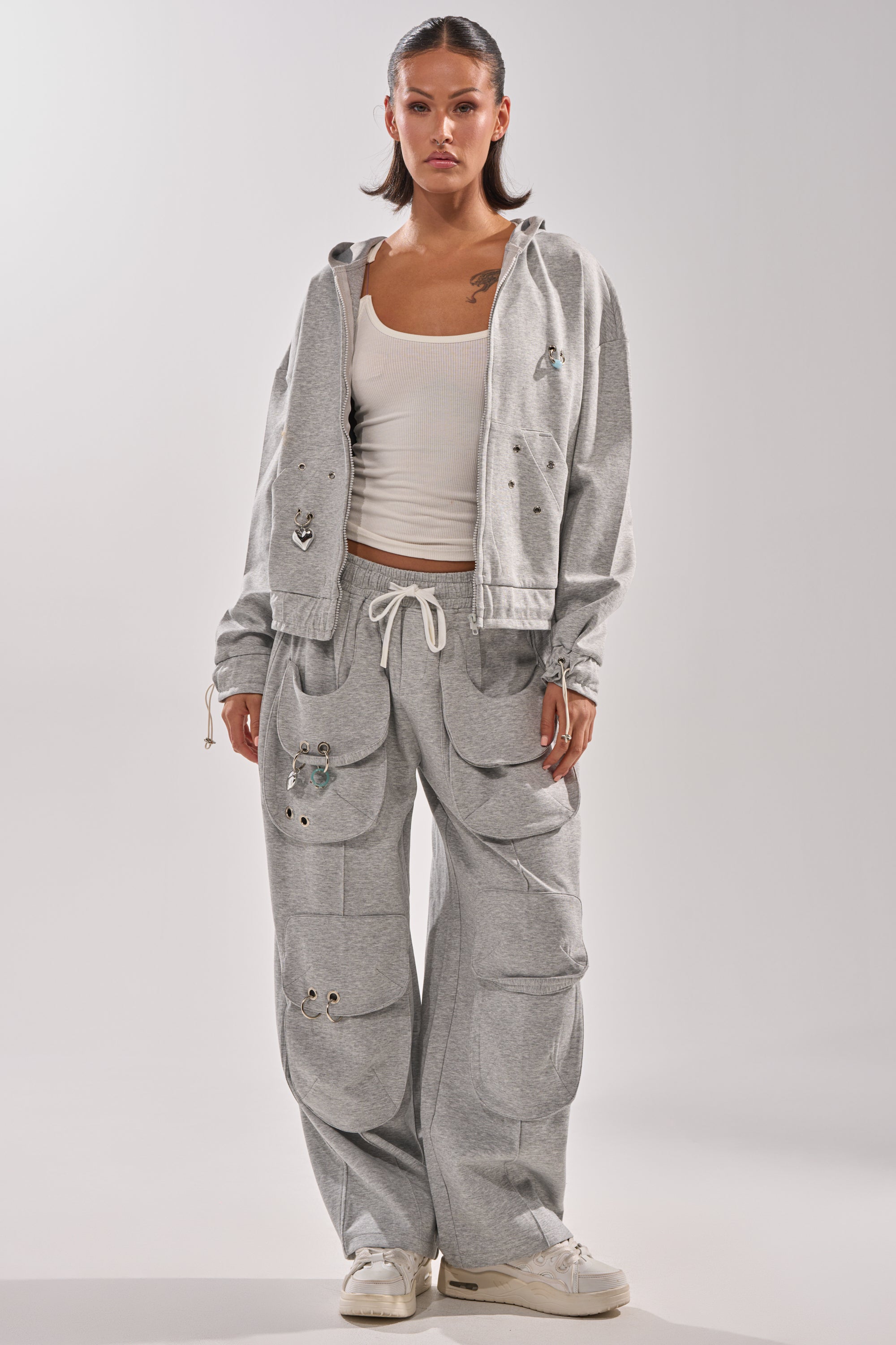 A woman faces forward in the GET WITH IT ZIP UP SWEATSHIRT, a light gray streetwear hoodie with multiple large patch pockets, paired with matching wide-leg sweatpants, a white tank top, and white sneakers against a plain background. 7