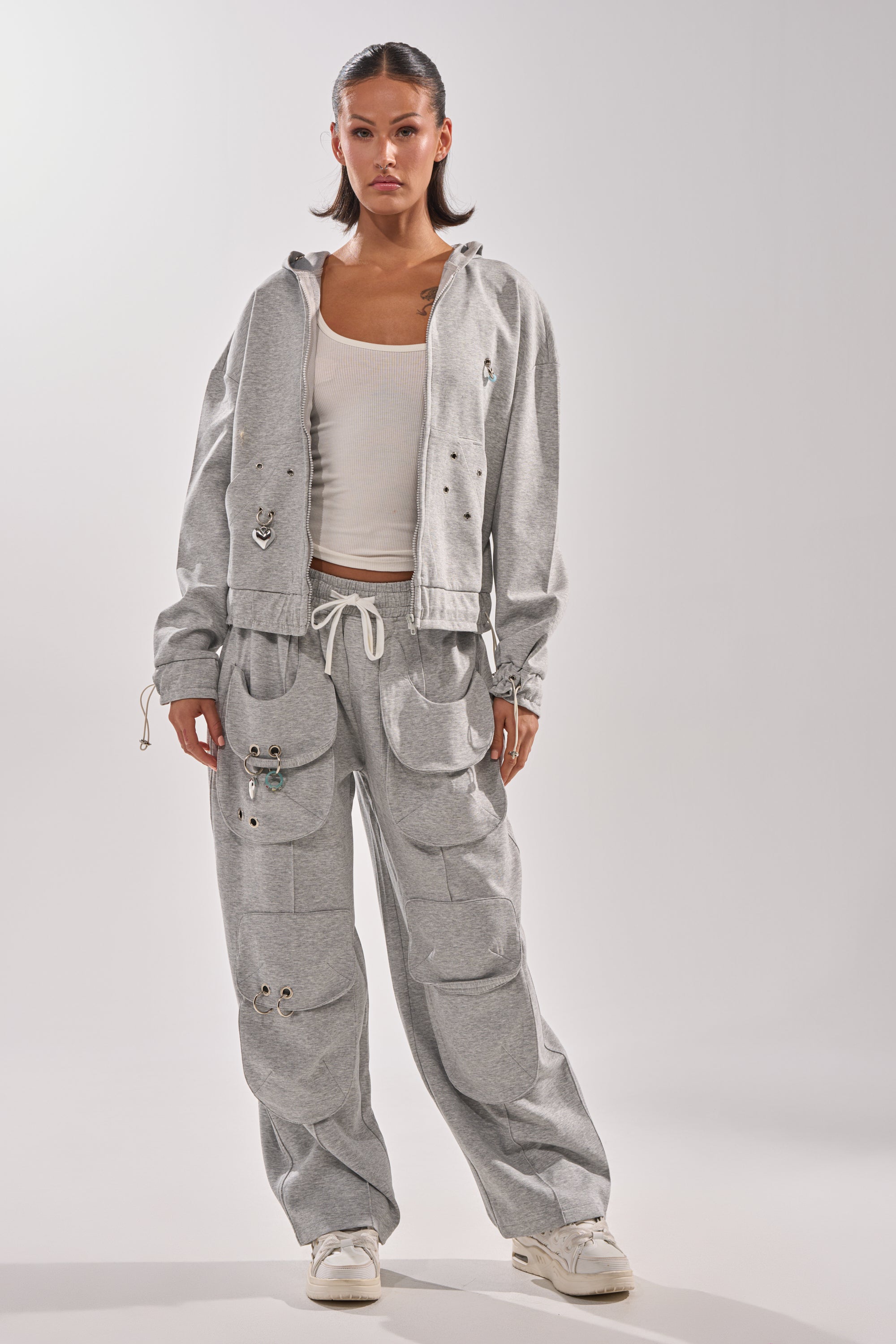 A woman with dark hair pulled back and a neutral expression faces forward, wearing the GET WITH IT ZIP UP SWEATSHIRT in light gray streetwear style with multiple pockets and metal eyelets, paired with a white tank top and white sneakers. 8