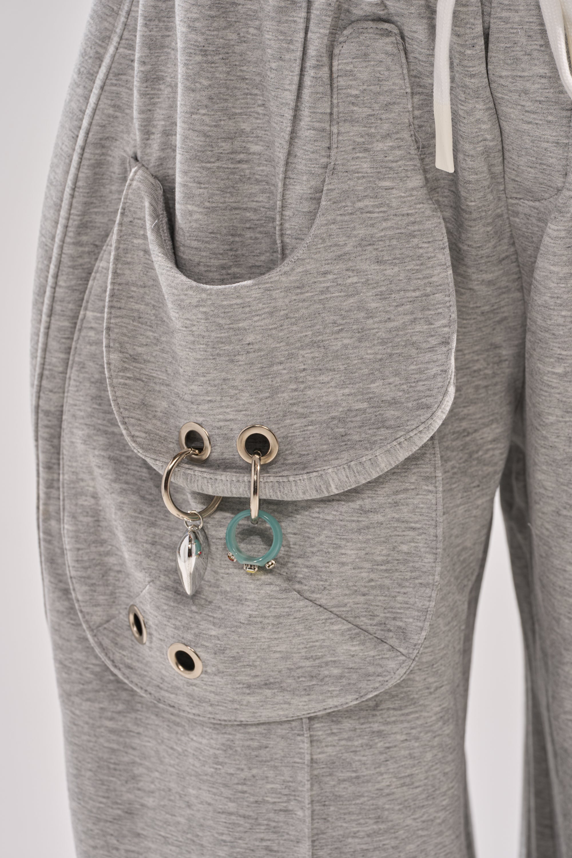 Close-up of the GET WITH IT WIDE LEG CARGO JOGGER in grey fleece, showcasing a large rounded pocket with metal grommets, decorative keychains (teal ring and silver charm), and white drawstrings for a modern streetwear look. 6