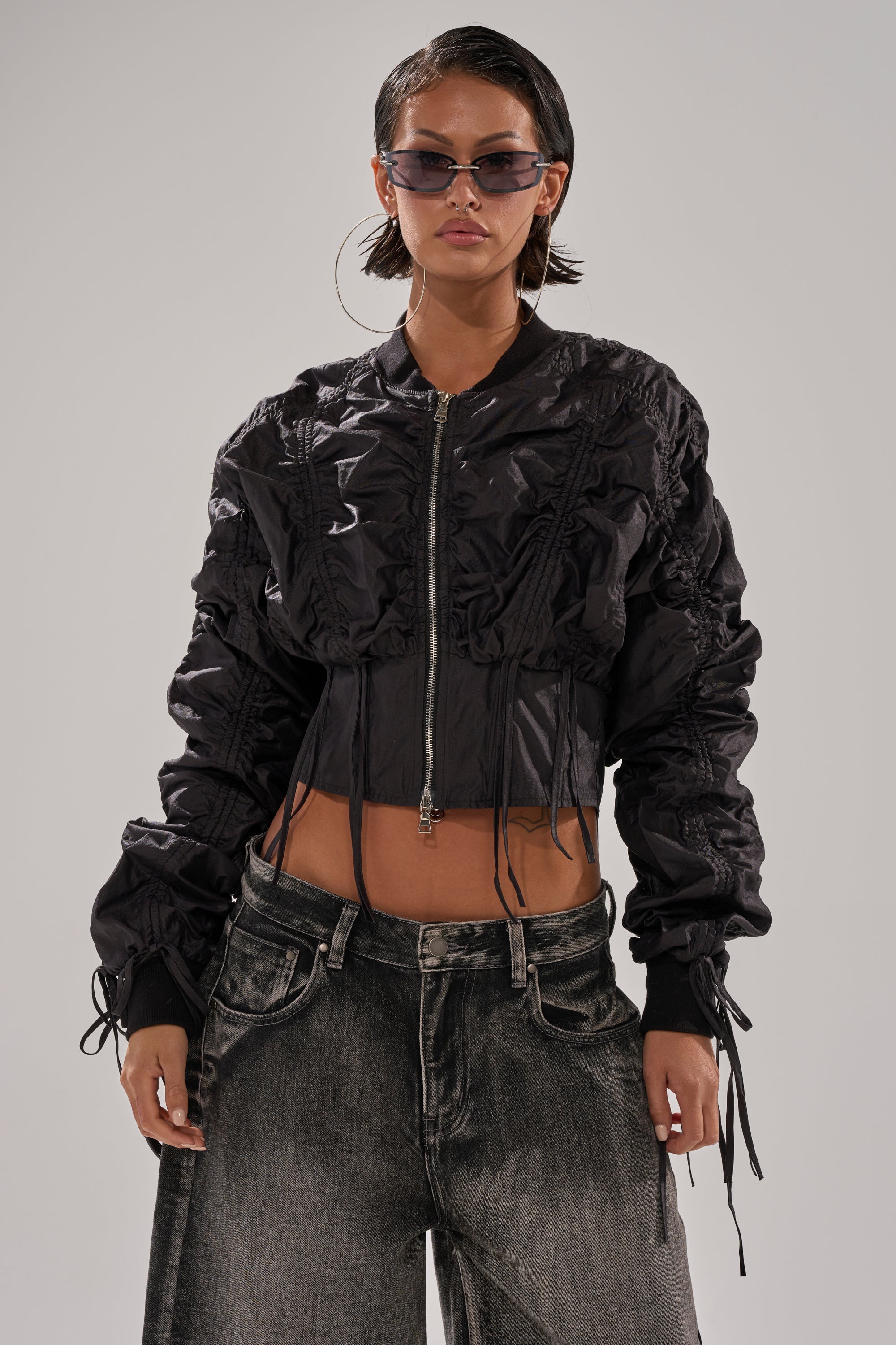 A person with short dark hair wears sunglasses, large hoop earrings, the ARIANA SCRUNCH BOMBER in shiny black with ruched details, and loose-fitting faded black jeans against a neutral gray background. 1