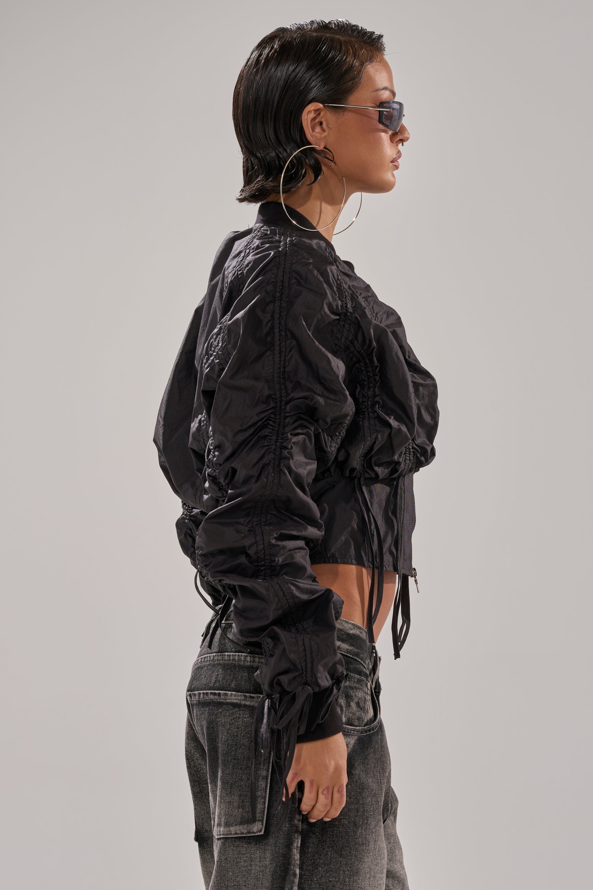 A woman with slicked-back hair wears hoop earrings, sunglasses, the ARIANA SCRUNCH BOMBER styled as a cropped black jacket, and faded gray jeans while standing in profile against a plain gray background. 2