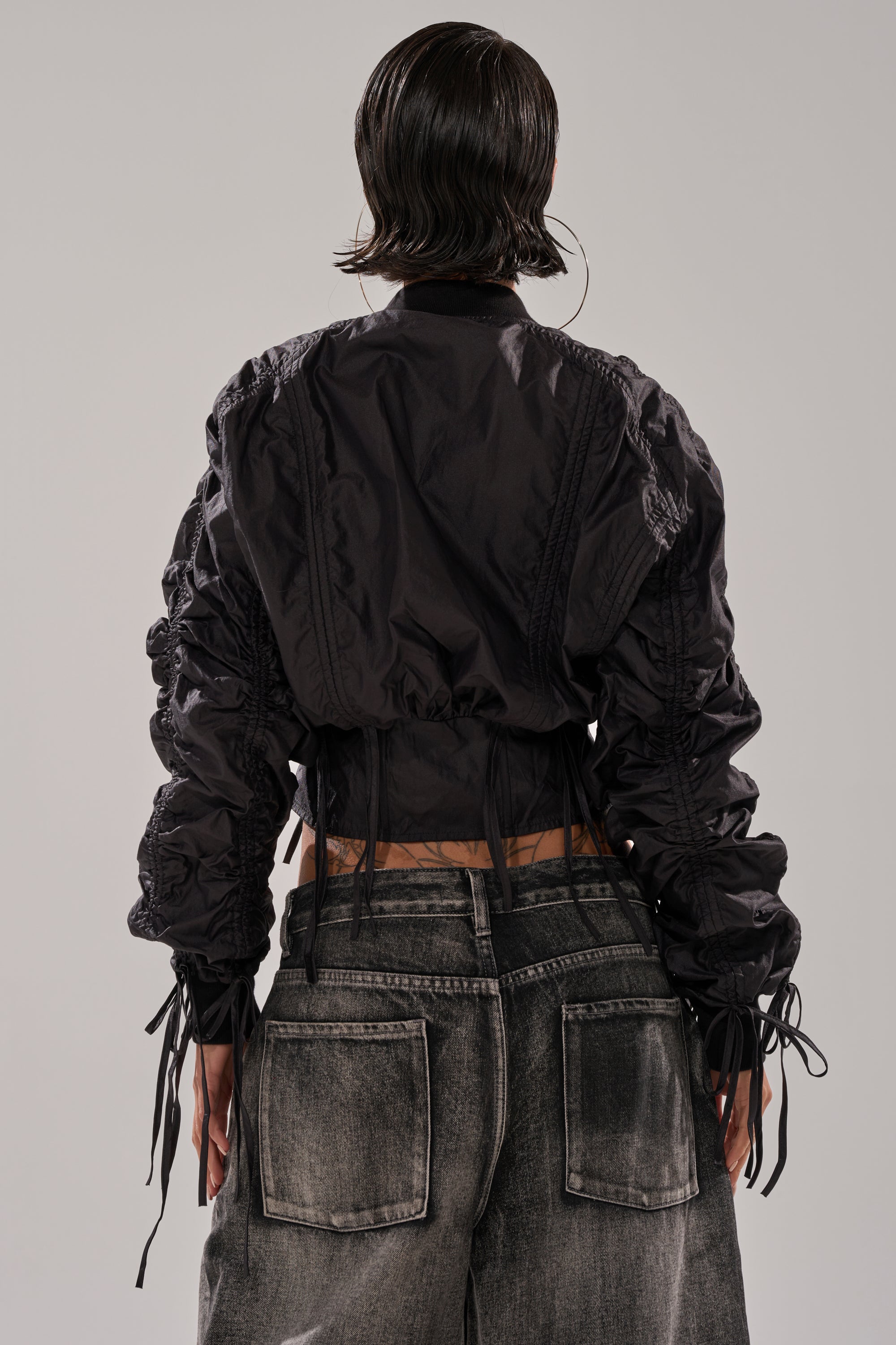 A person with short, wet-look hair is seen from behind, wearing the ARIANA SCRUNCH BOMBER and loose-fitting, washed gray denim jeans with visible stitching and back pockets. 3
