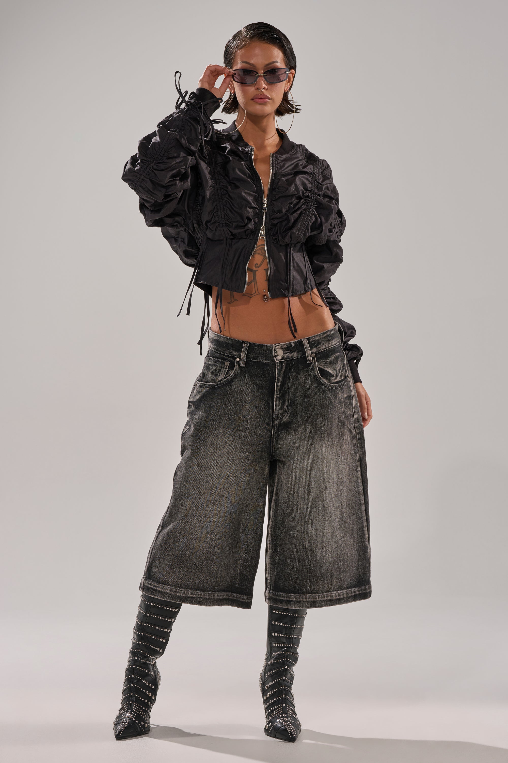 Wearing the ARIANA SCRUNCH BOMBER with wide-leg faded denim shorts, sheer black striped knee-high socks, sunglasses, and large earrings, a person poses confidently against a plain light background. 4