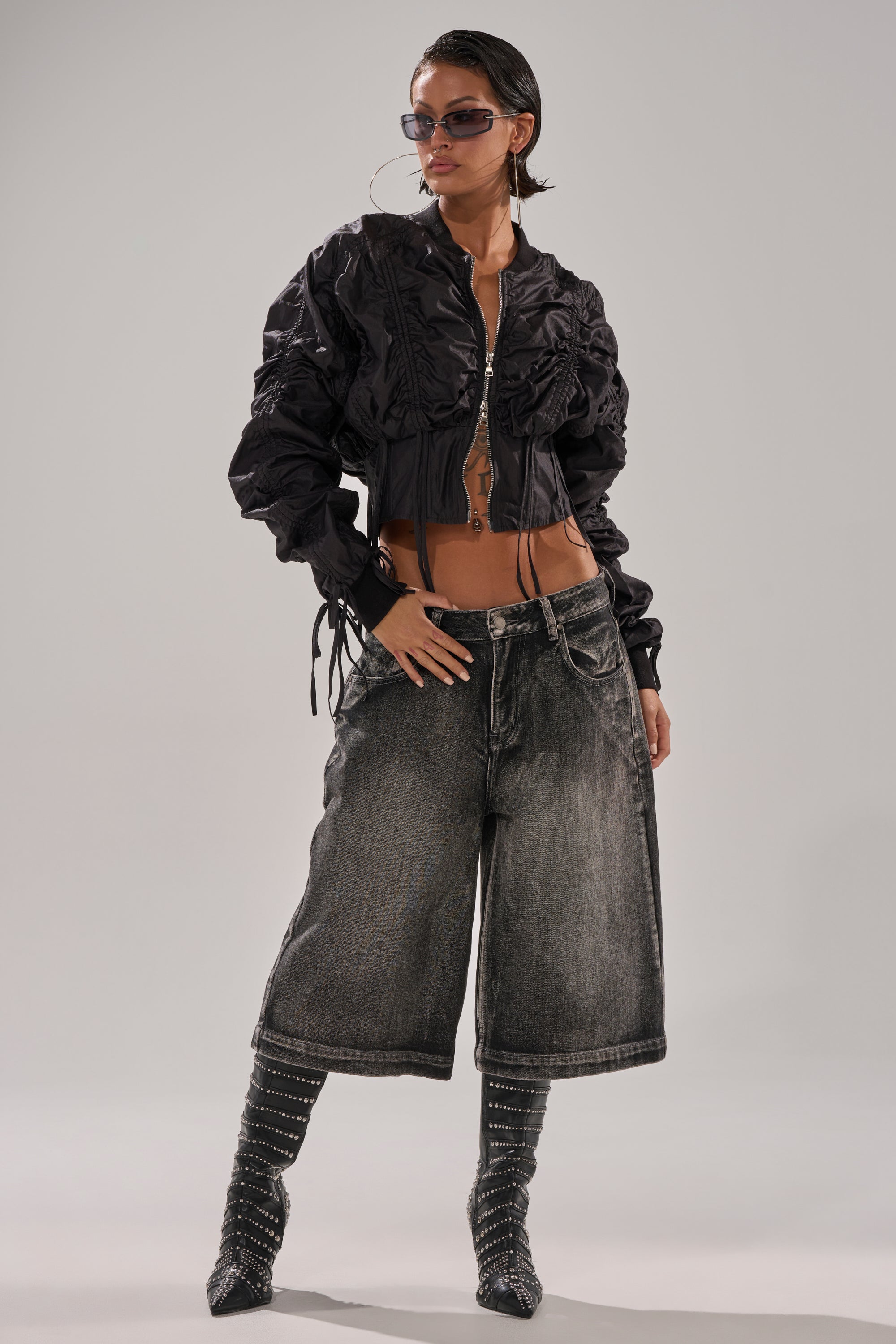 Wearing sunglasses, the model poses confidently in the ARIANA SCRUNCH BOMBER, black and ruched, styled with wide-leg faded denim shorts, hoop earrings, and dotted pointy-toed knee-high stockings against a plain background. 5