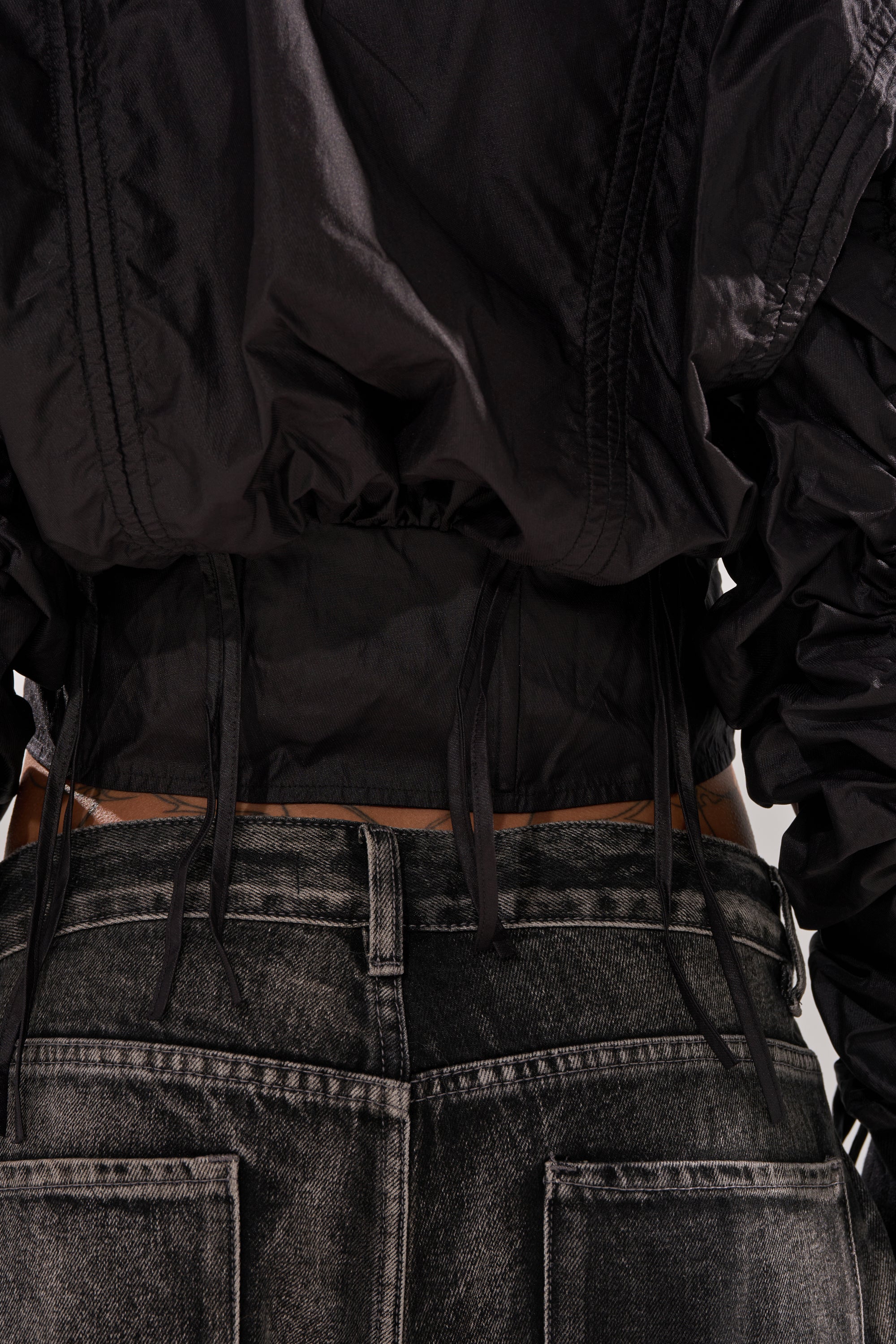 Close-up of a person’s lower back wearing the ARIANA SCRUNCH BOMBER layered over a black ruched top with ties, paired with faded black denim jeans featuring visible belt loops for an edgy look. 6