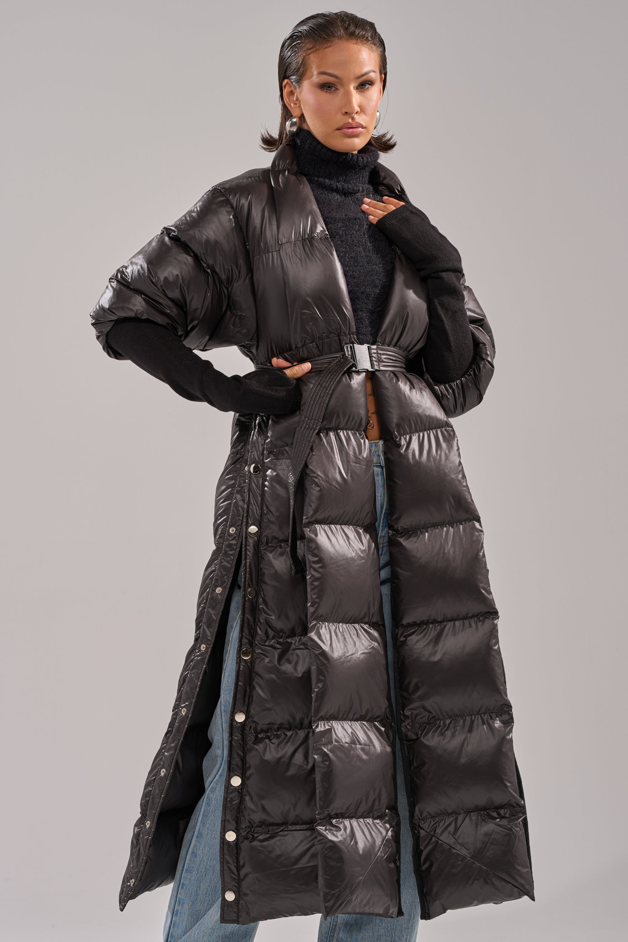 A woman models the POPPIN OFF TEARAWAY PUFFER—an elegant long black quilted coat with a belt—styled over a black turtleneck and blue jeans against a simple gray background.